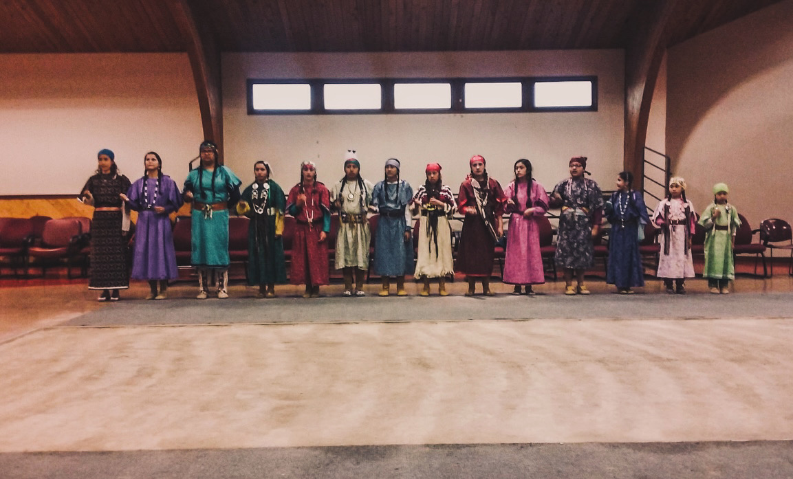 Group of people in traditional clothing standing in a line indoors, with windows and wooden walls behind them.