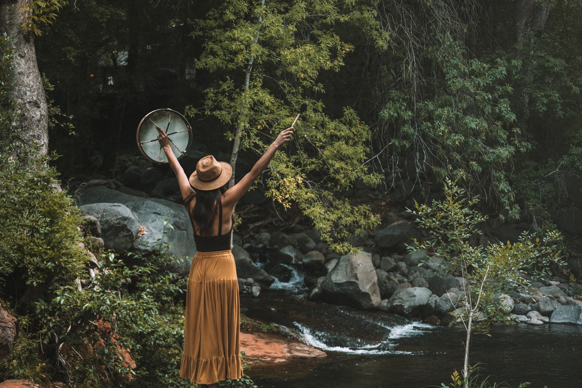 Woman with arms raised, wearing a hat and long skirt, standing near a river with trees and rocks in the background.
