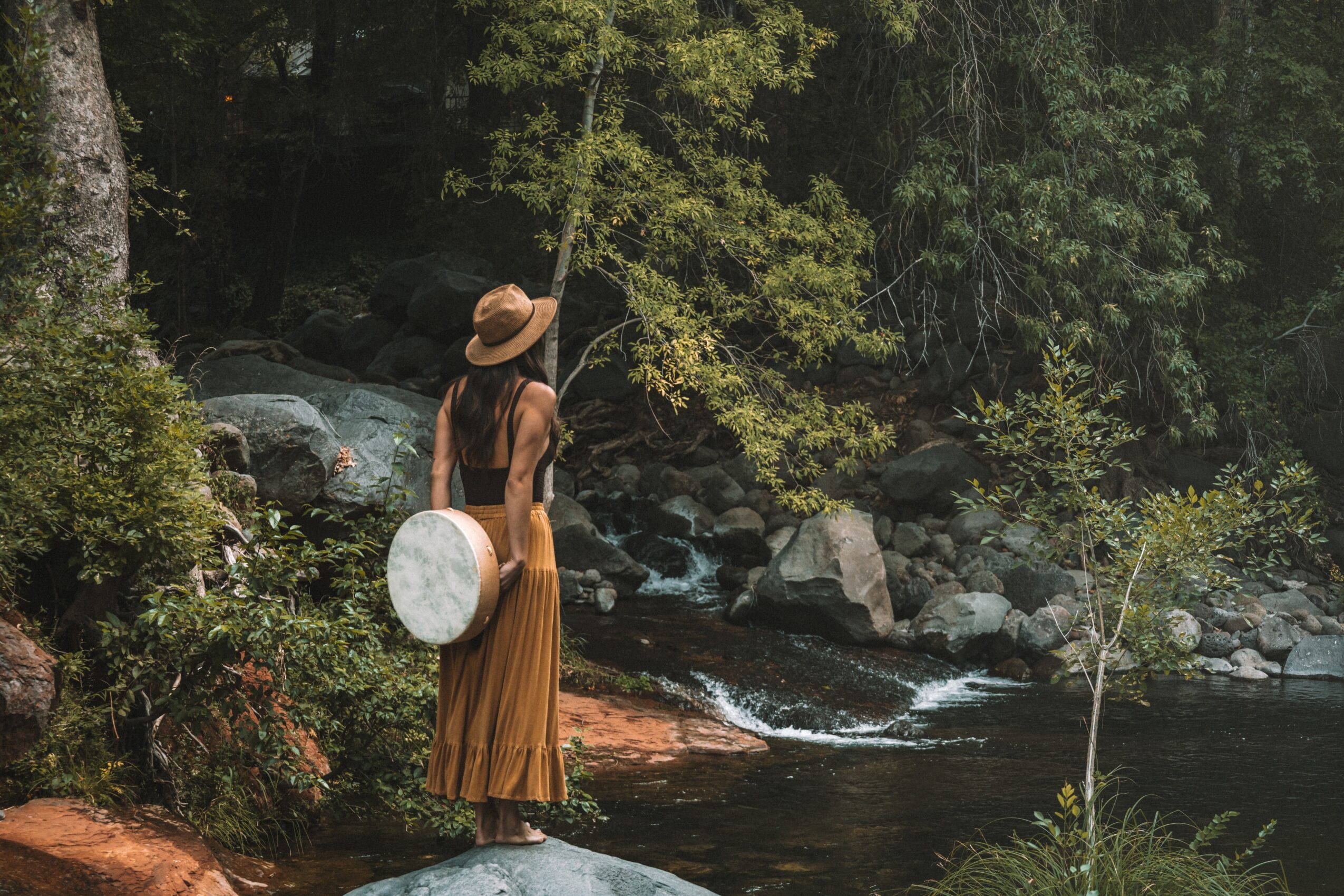 Woman in a long skirt and hat standing by a river in a forest, fishing with a rod, surrounded by trees and rocks.
