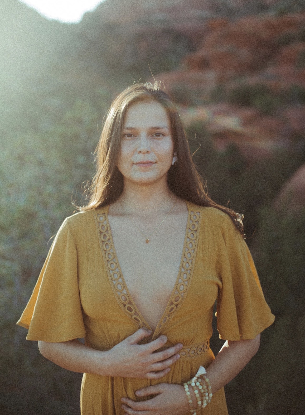 Young woman with long hair wearing a mustard yellow dress with a deep neckline, standing outdoors with a natural background.