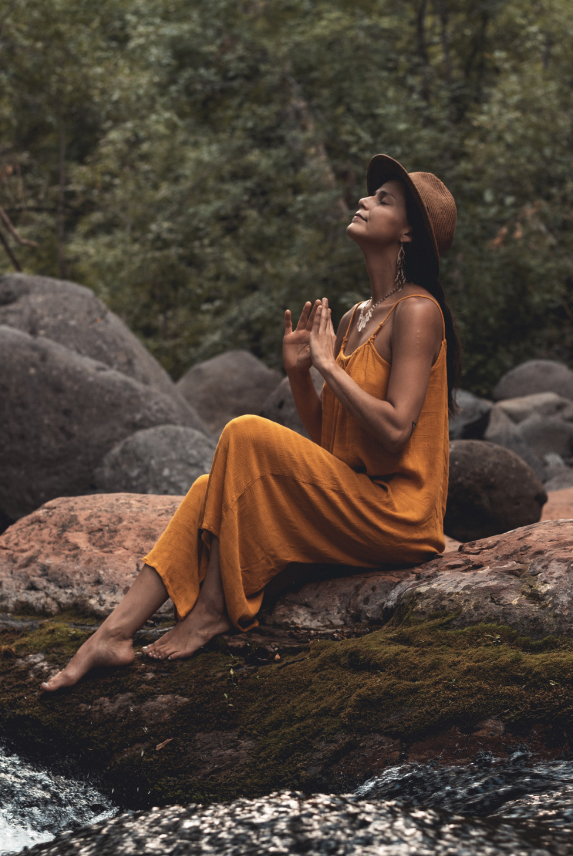 Woman in orange dress and hat sitting on rocks with hands in prayer position, surrounded by nature and water.