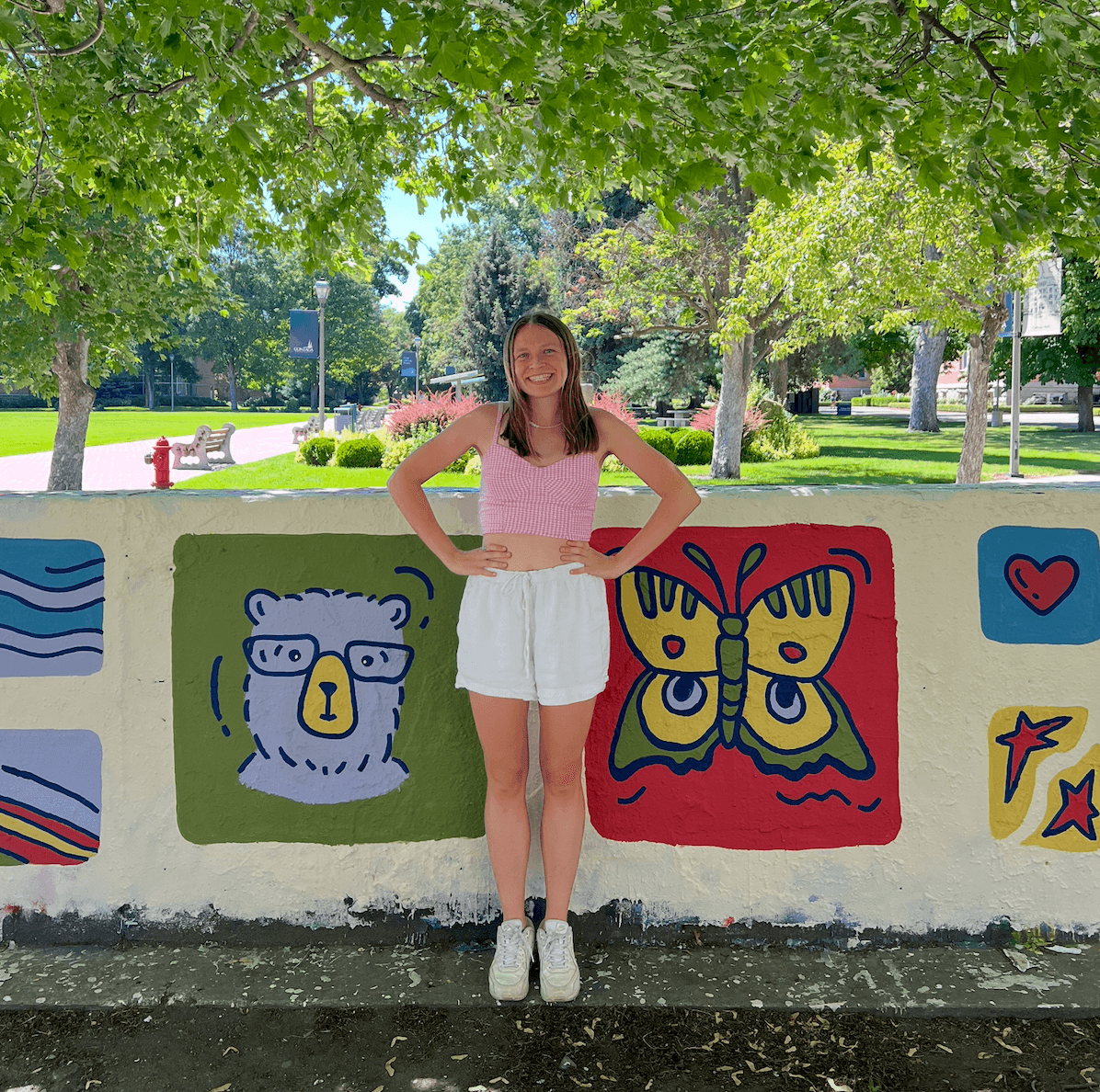 Young woman standing outdoors in front of colorful painted wall, smiling, wearing pink top and white shorts, trees and park in background.