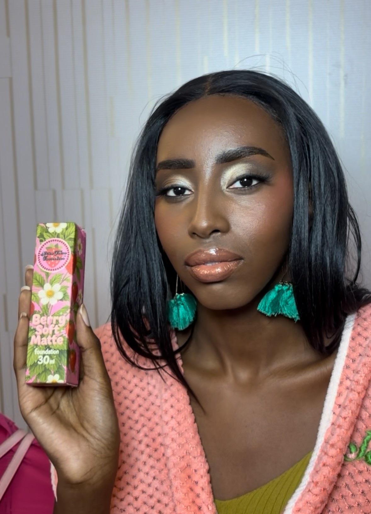 Woman with black hair and turquoise earrings holding a pink and green box, standing in front of a light-colored background.