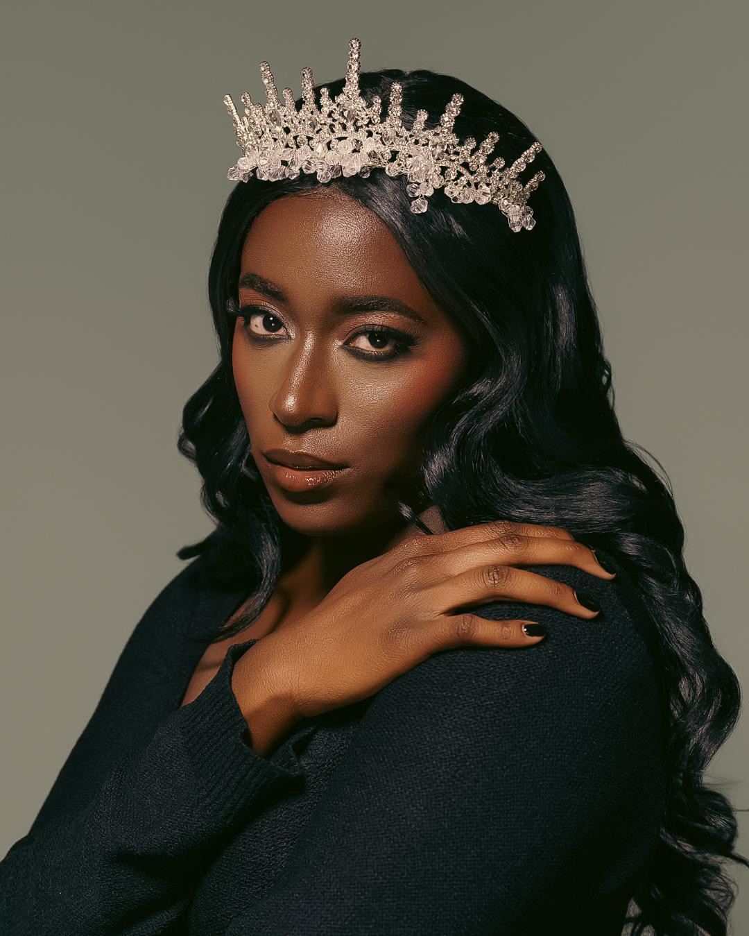 Woman with long dark hair wearing a crown, looking at the camera, hand near face, neutral background.