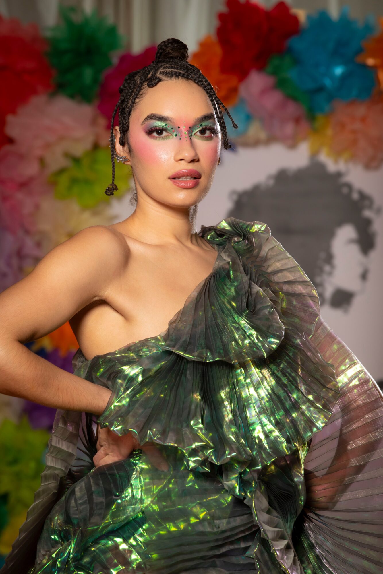 Young woman with braided hair and colorful makeup, wearing a shiny green dress, standing in front of colorful paper flowers.