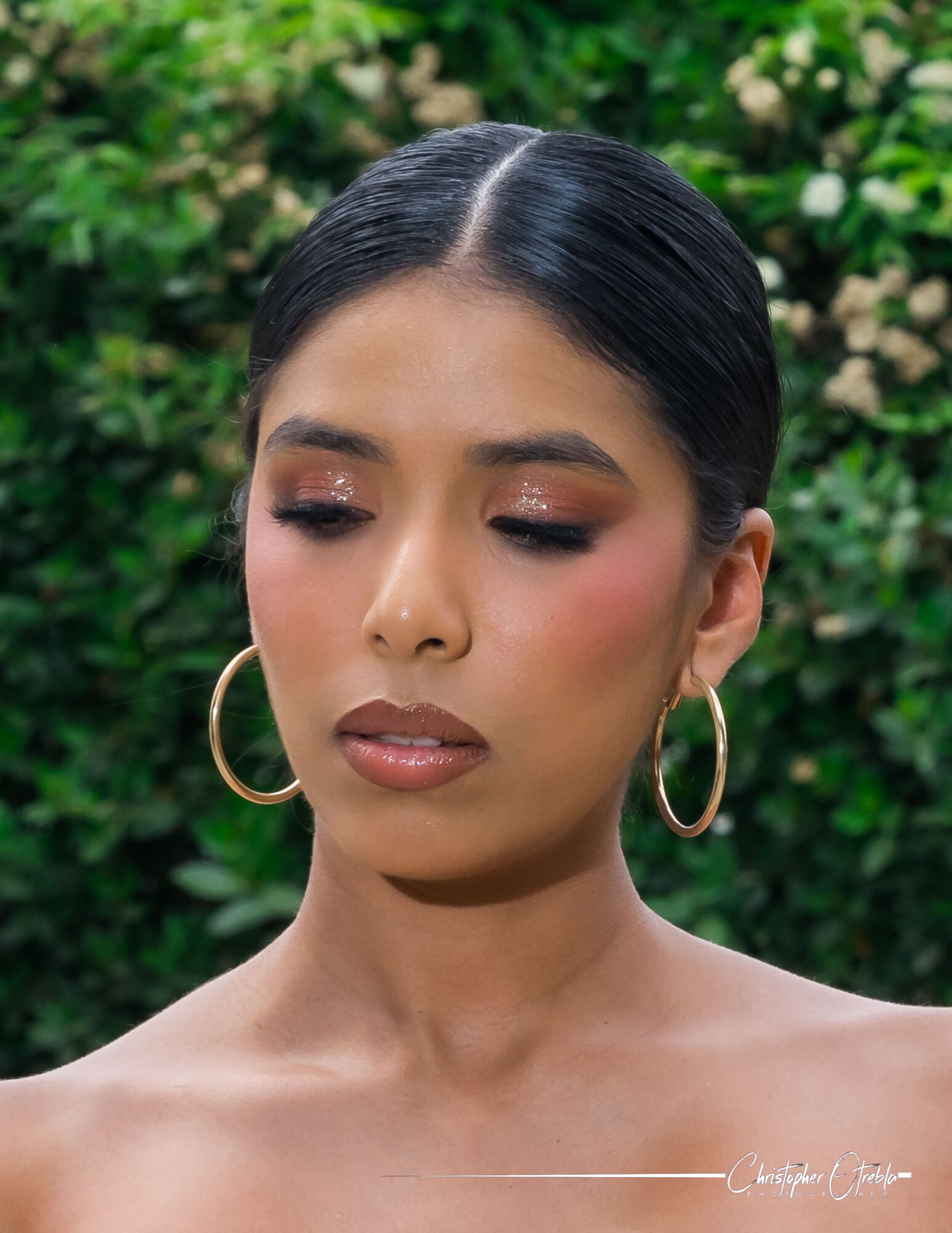 Portrait of a woman with dark hair in a bun, wearing large hoop earrings, with a green leafy background.