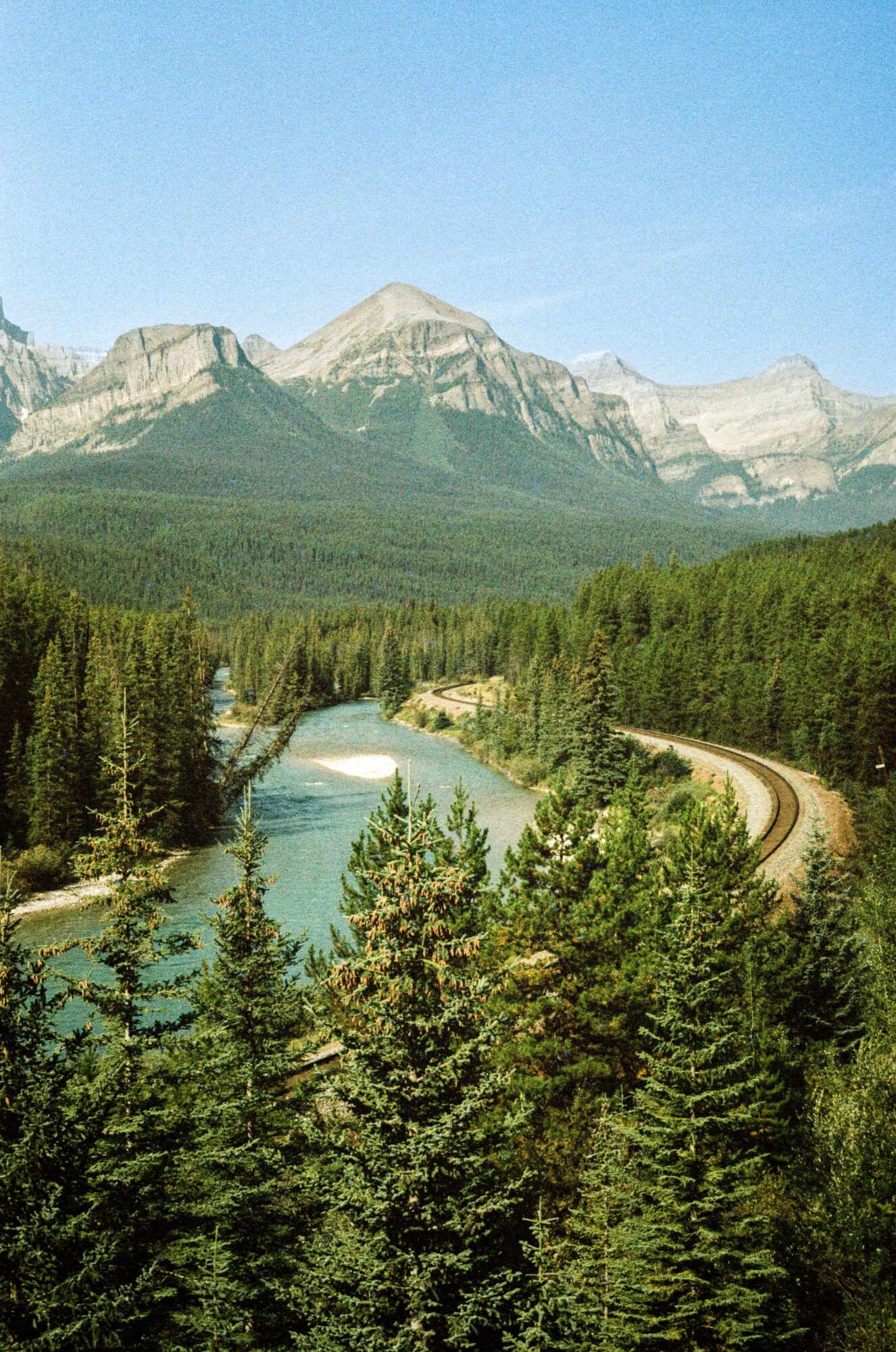 Mountain range with forested foreground, river, and a winding road, under a clear blue sky.