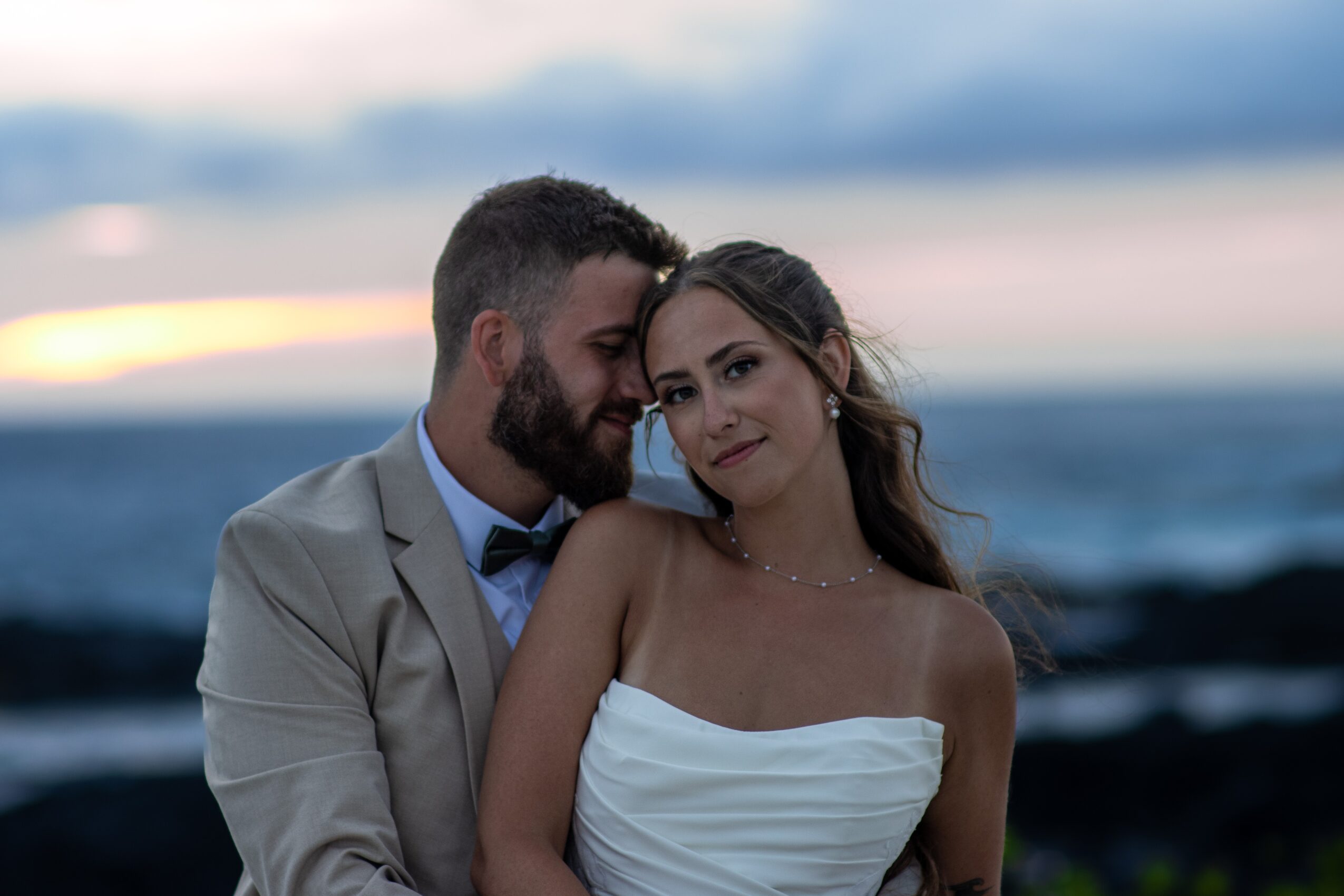 A couple stands close together outdoors at sunset, with the ocean in the background, smiling softly.
