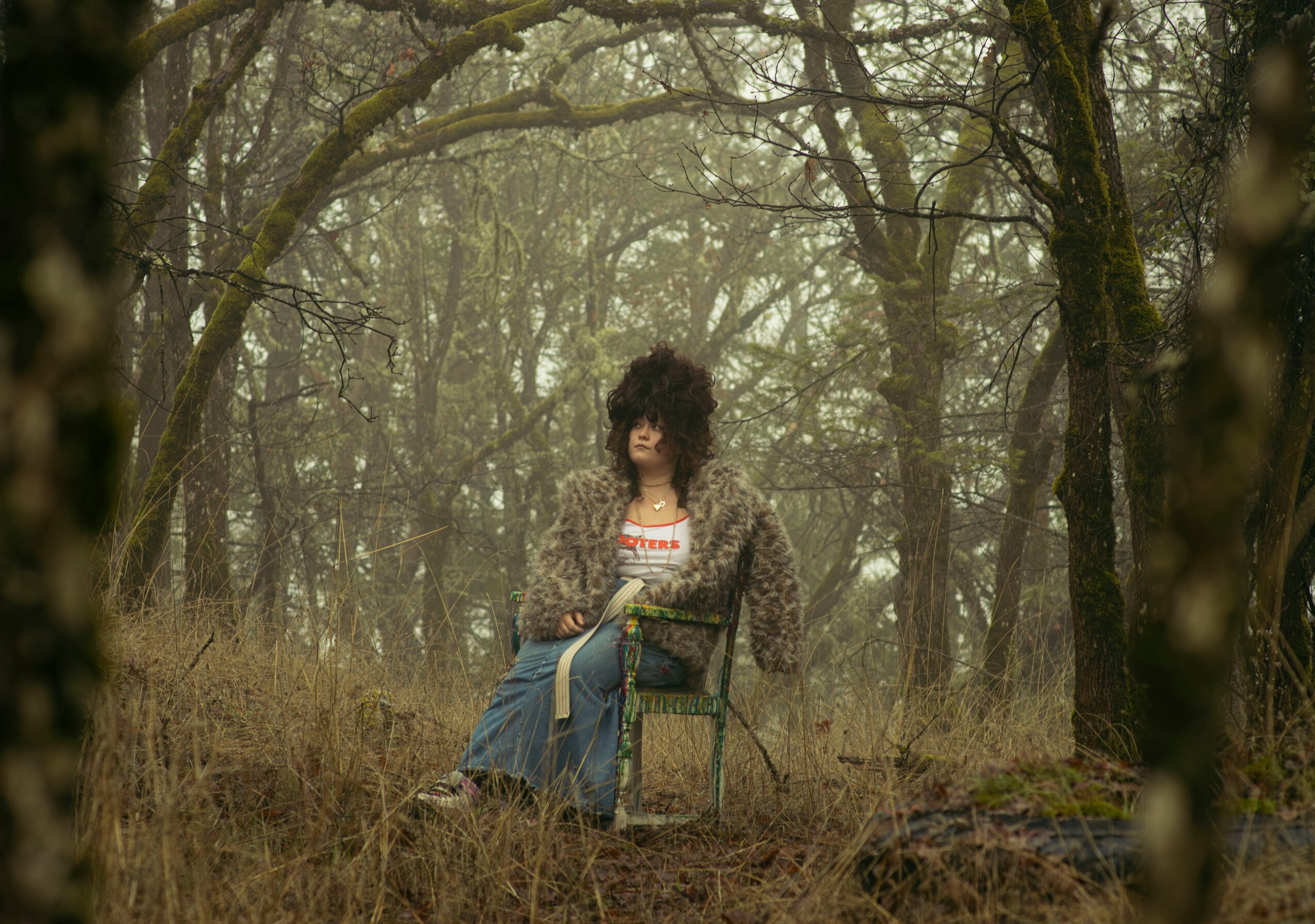 Person with curly hair sitting on a chair in a forest, surrounded by trees and foliage.
