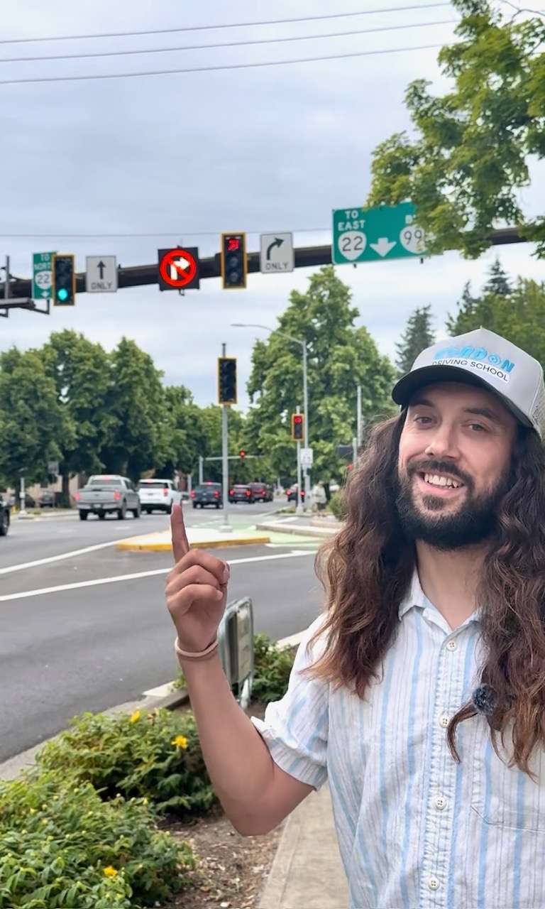 Smiling man with long hair and beard pointing upward outdoors near traffic lights and trees.