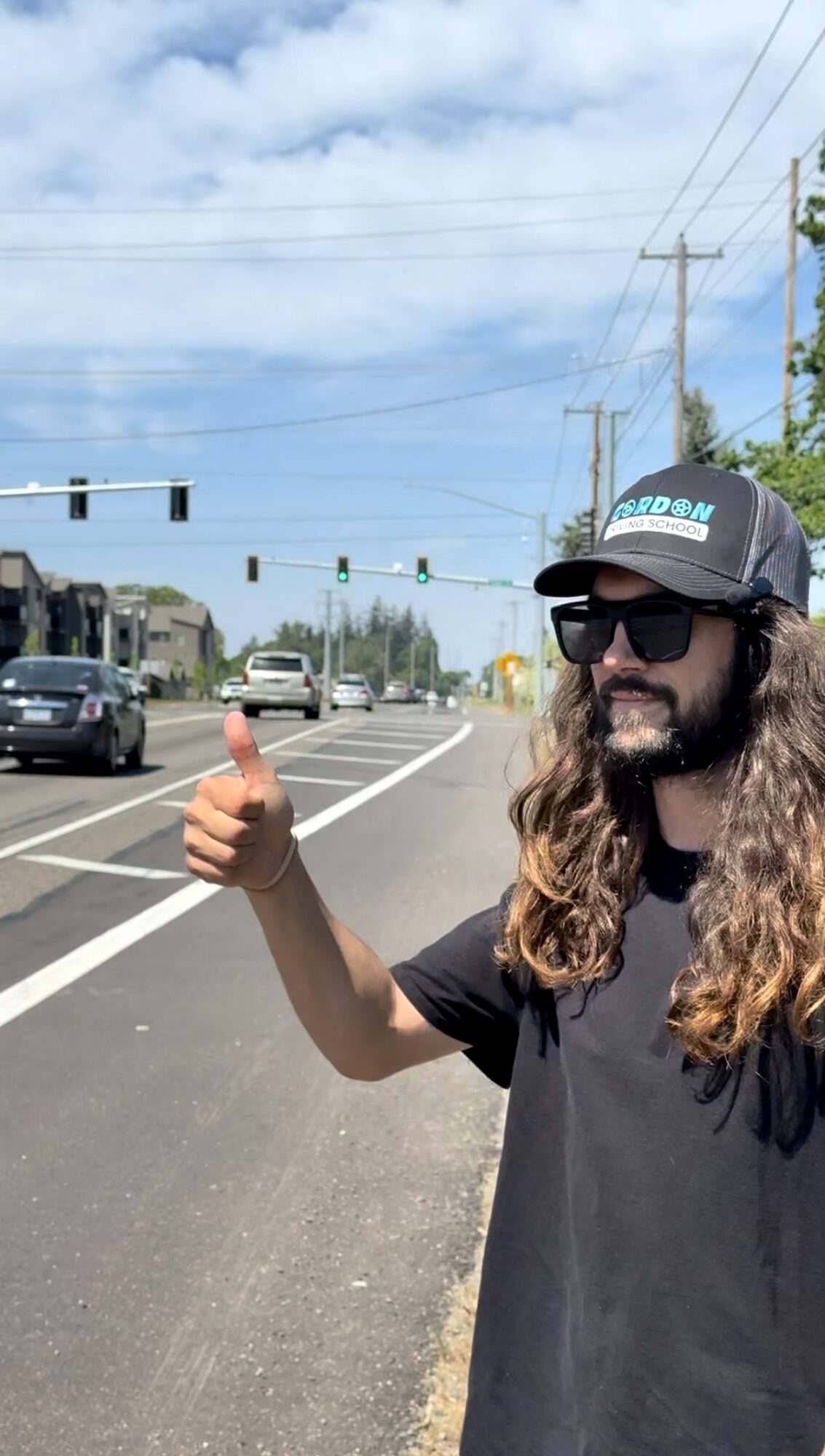 Person with long hair, sunglasses, and a cap giving a thumbs-up on a street with traffic lights and cars.