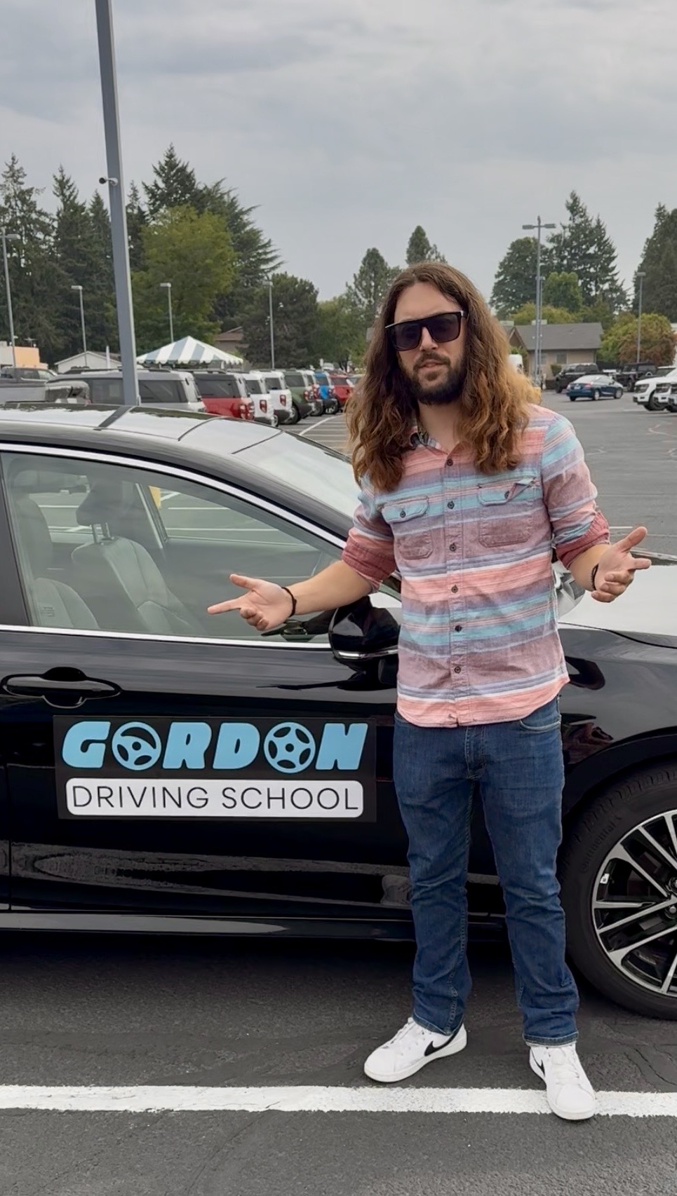 Person with long hair and sunglasses standing next to a car with a 'Gordon Driving School' sign, outdoors in a parking lot.
