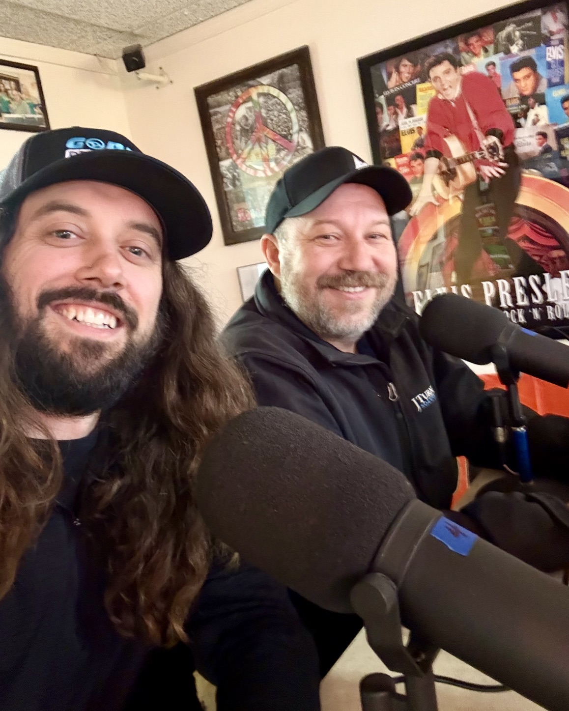 Two men with beards and caps smiling at a microphone in a room with framed posters on the wall.