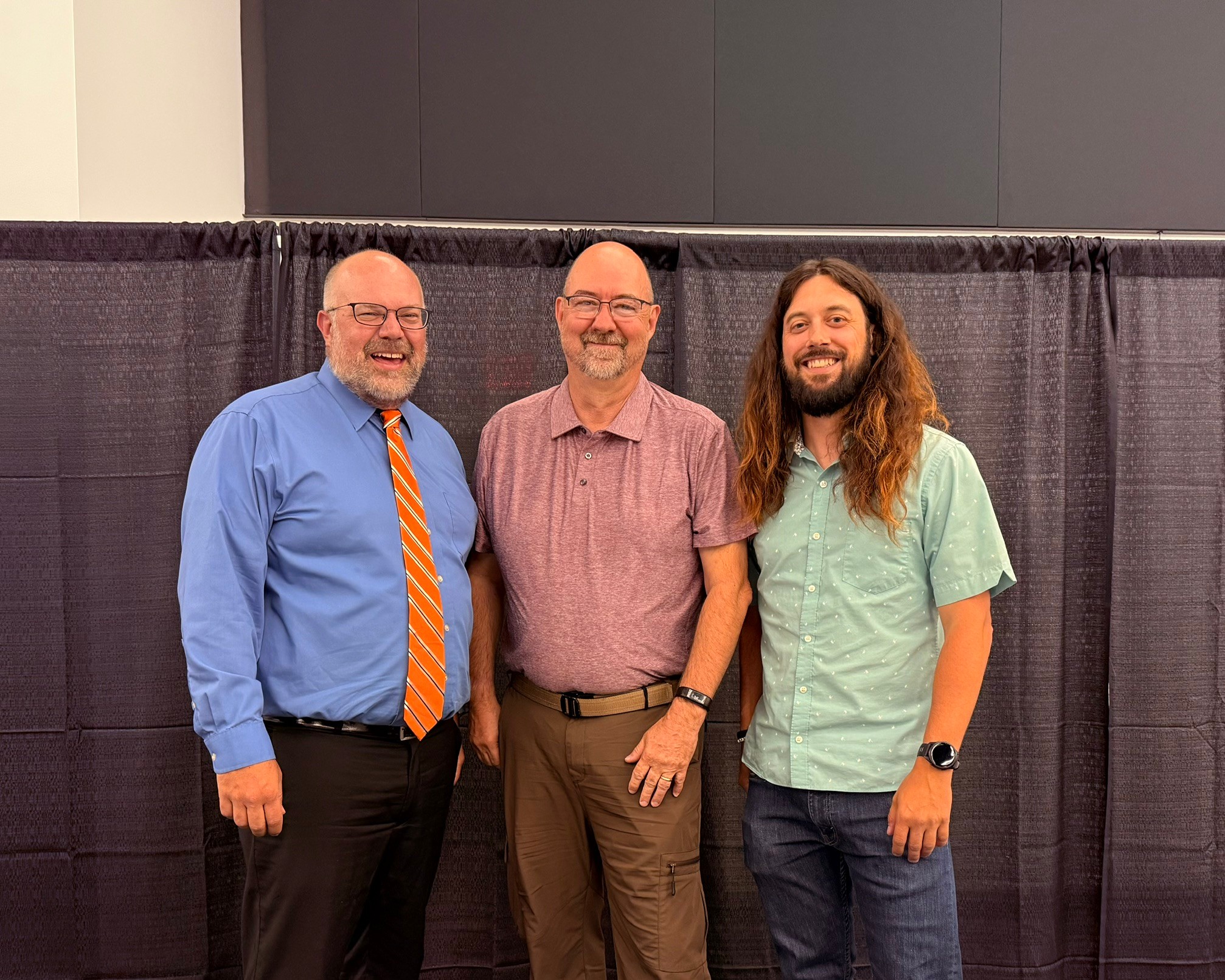 Three men standing side by side in front of a black curtain, smiling, indoors.