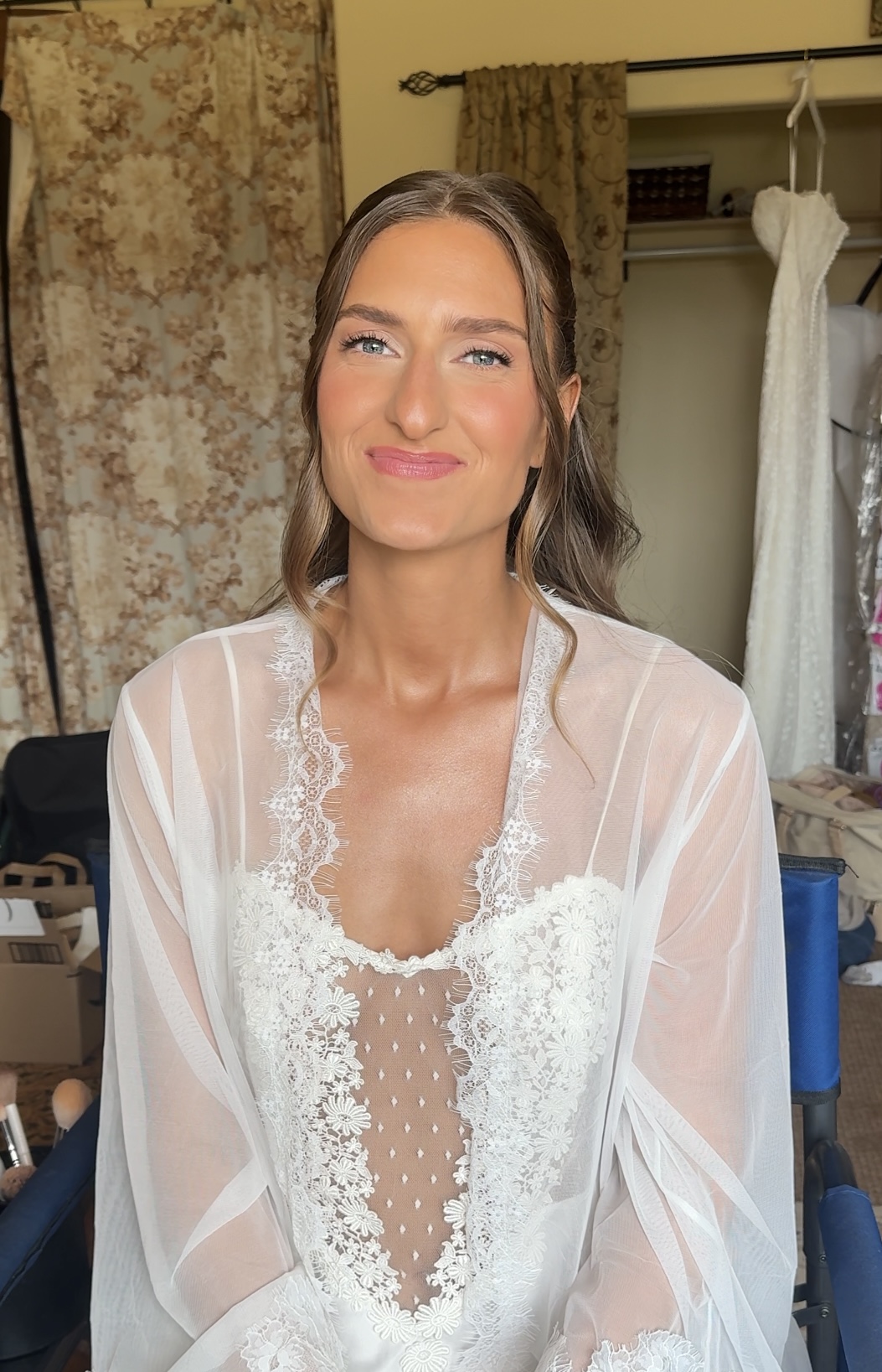 Young woman with long hair wearing a sheer white robe over a lace dress, smiling, in a room with curtains and clothing racks.