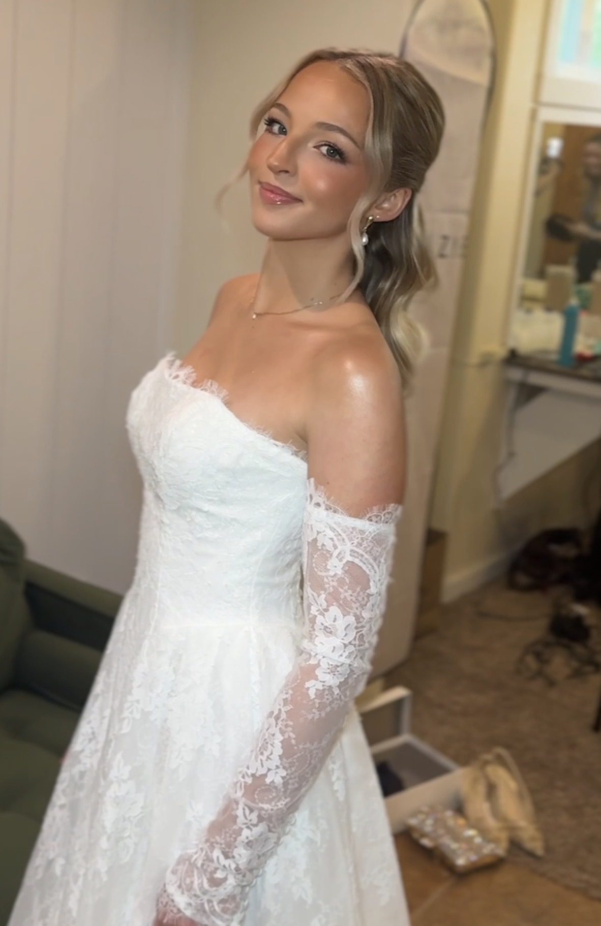 Young woman in a white lace off-shoulder dress, smiling, with blonde hair styled in loose waves, indoors.