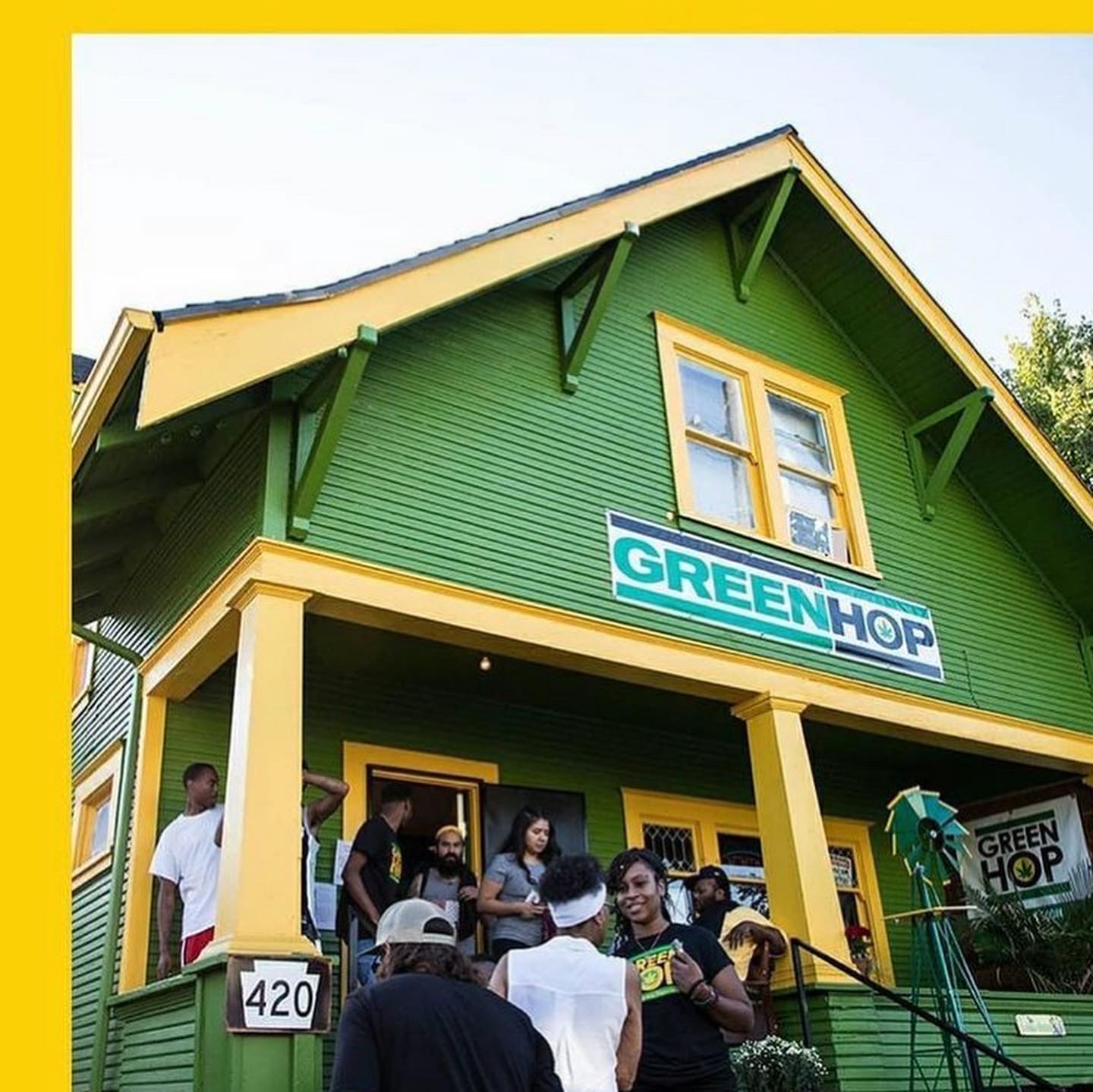 Green building with a group of people on the porch, sign reads 'GREEN HOP', yellow trim, blue sky.