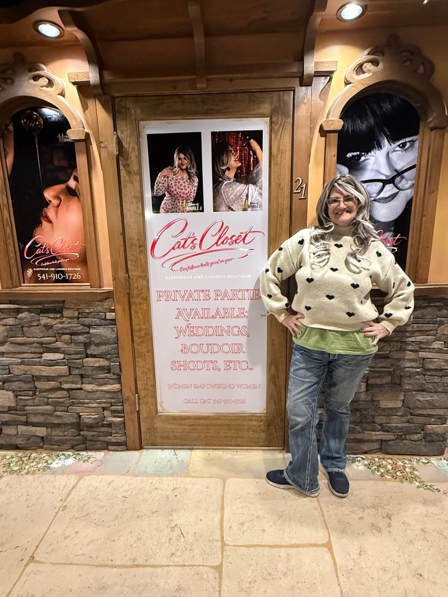 Person standing next to a wooden display board with photos and text, in front of a stone wall, wearing a patterned jacket and jeans.