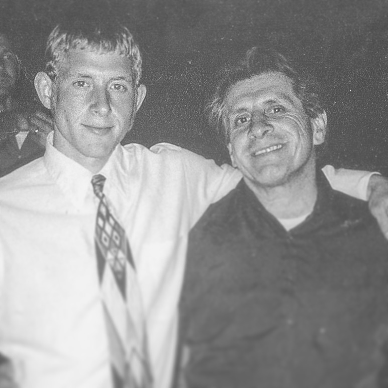 Two men smiling, one with arm around the other, in a black and white photo.