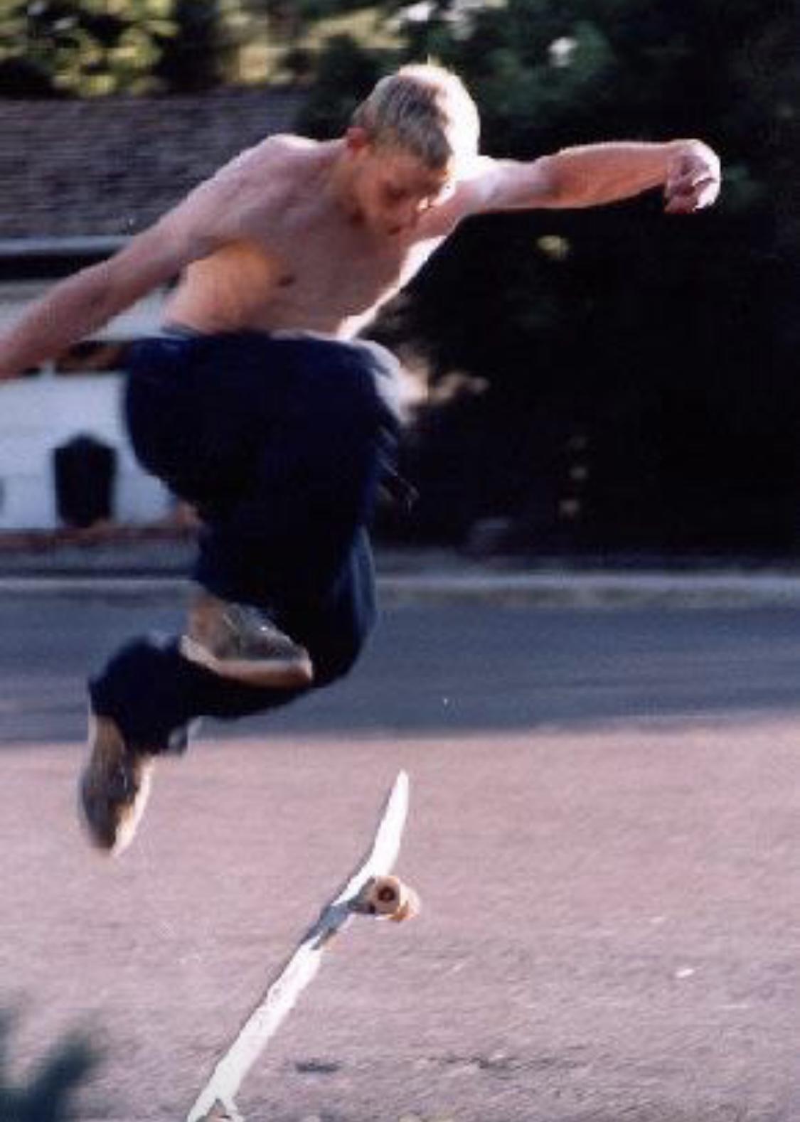 Young man shirtless, mid-air performing a skateboard trick outdoors, with trees and a building in background.