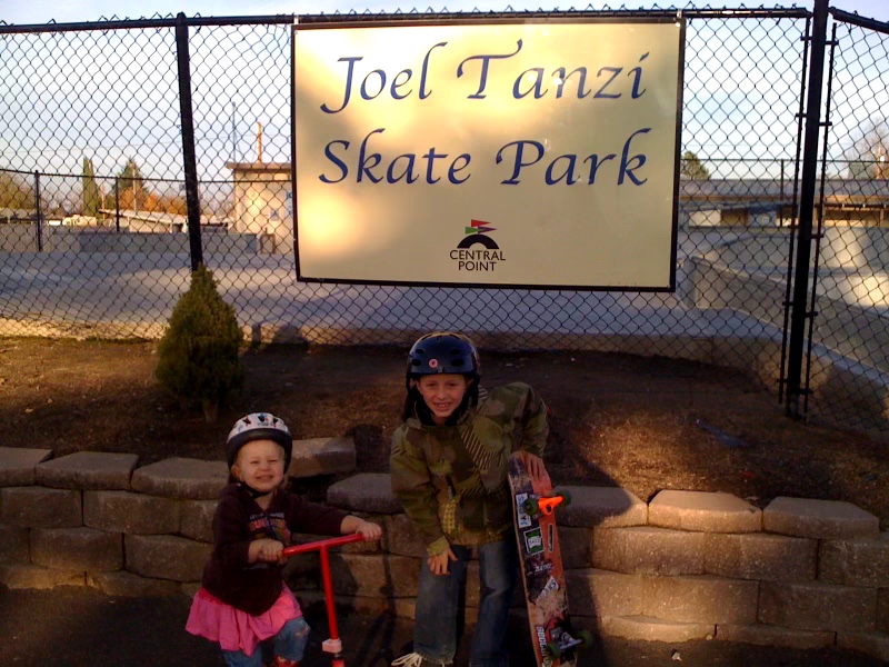 Two children with helmets and a woman pose with skateboards in front of a sign for Joel Tanzi Skate Park, with a fence and trees behind.