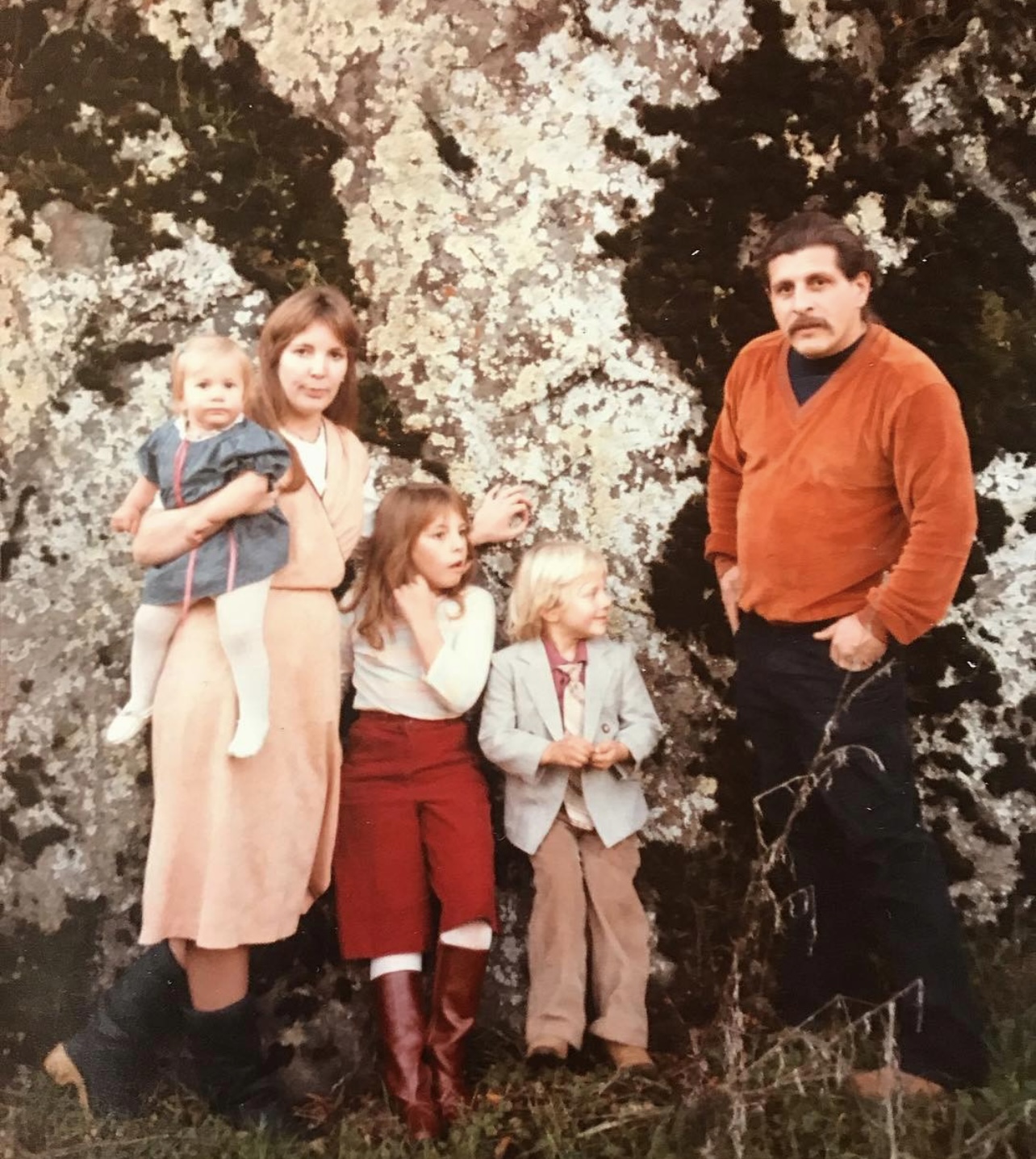 Group of five people standing outdoors in front of flowering trees, with children and adults, some holding objects.