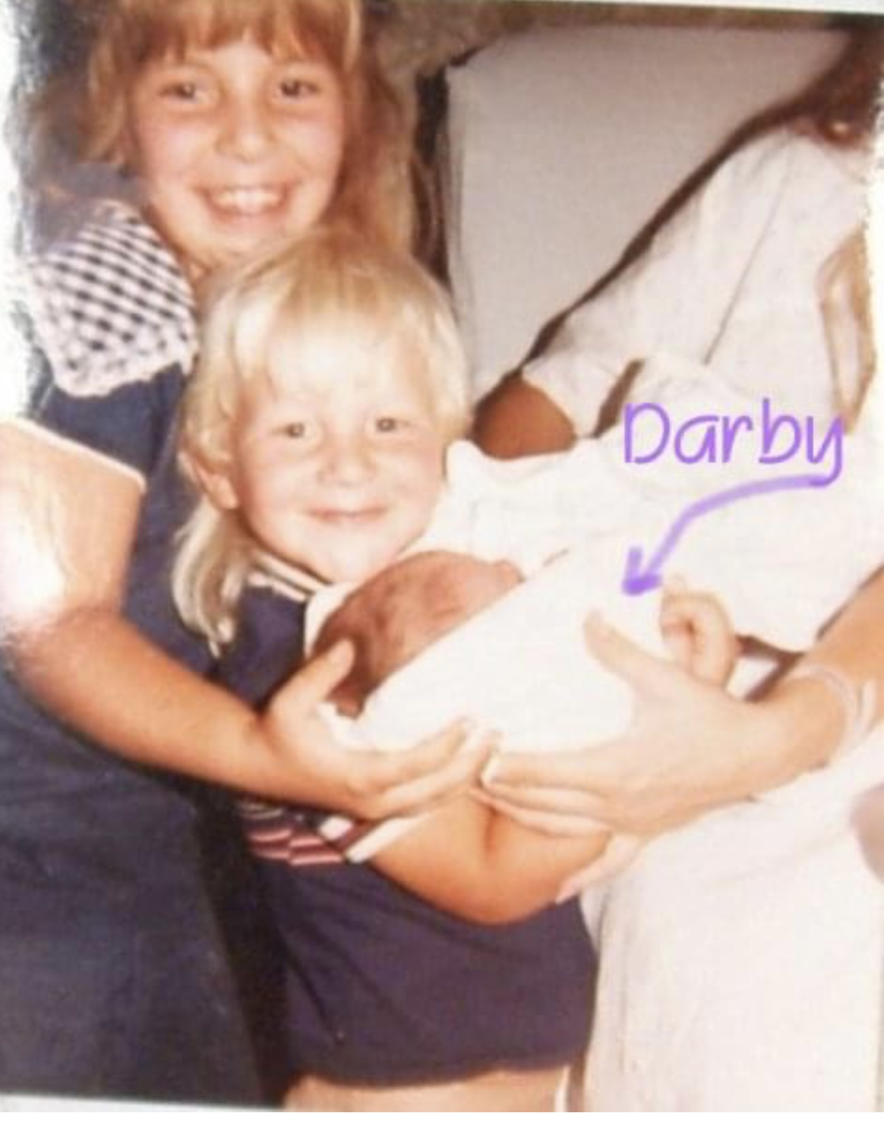 Two children smiling, holding a baby, with a person in white clothing nearby. The scene appears to be indoors.