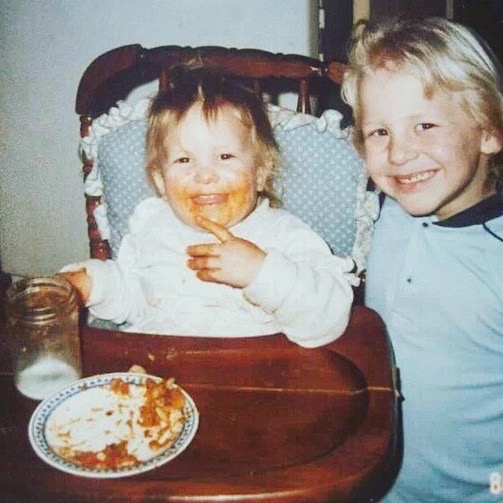 Two children smiling, one with food on face, sitting at a table with a plate of food, in a high chair and standing.