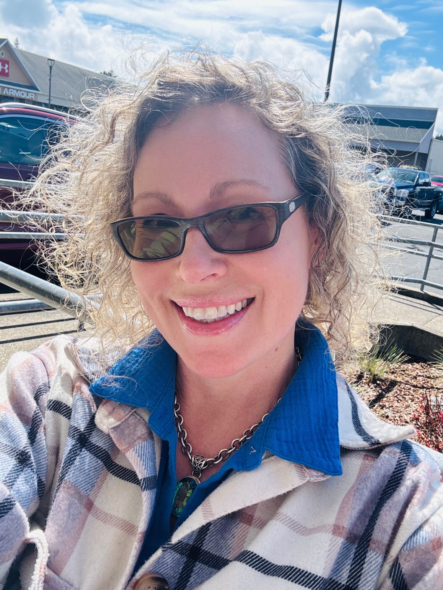 Person wearing sunglasses and a plaid jacket, smiling outdoors near parked cars.