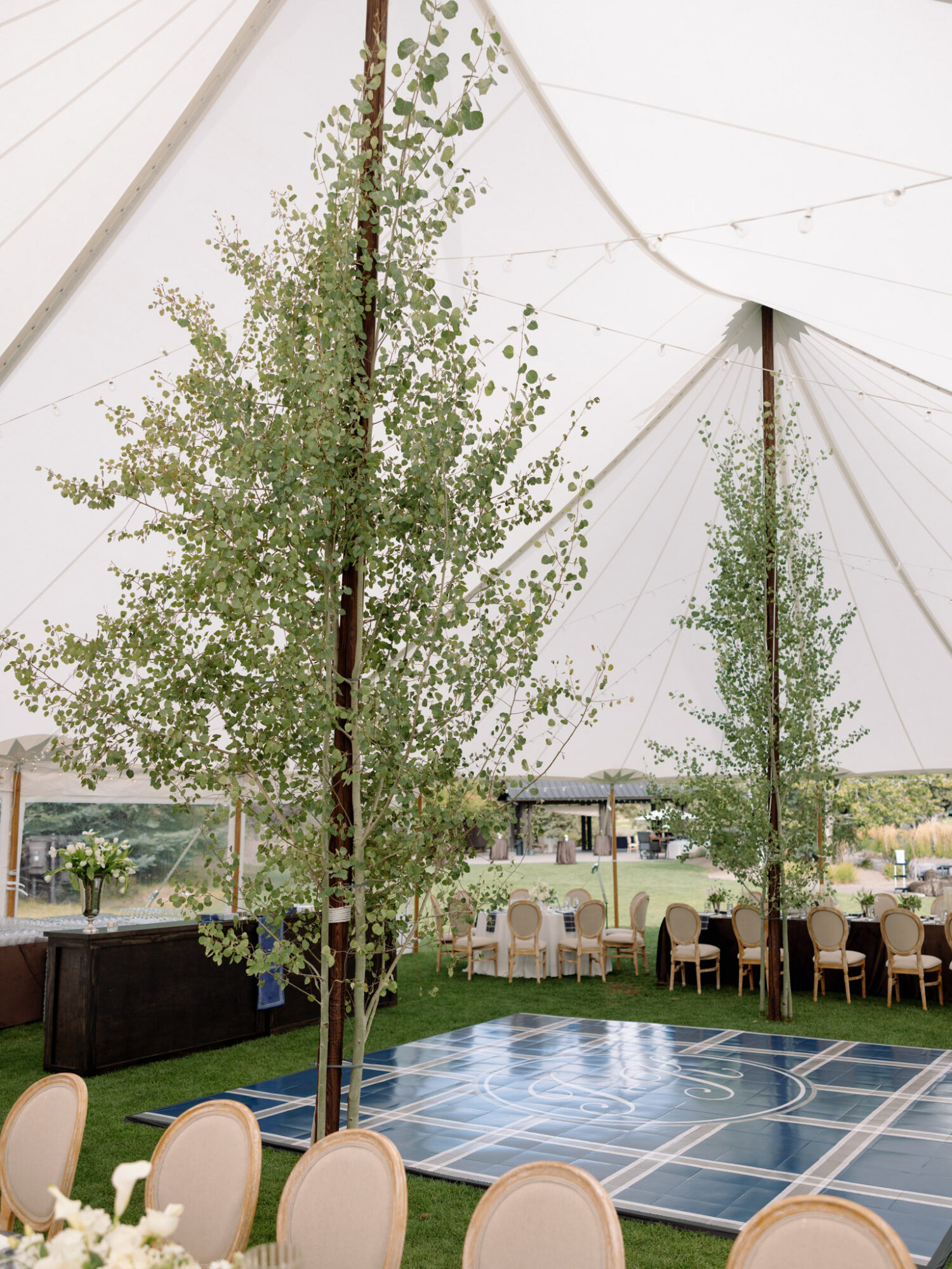 Event tent interior with trees, round tables, chairs, and a blue sports court, under a white canopy with supporting poles.