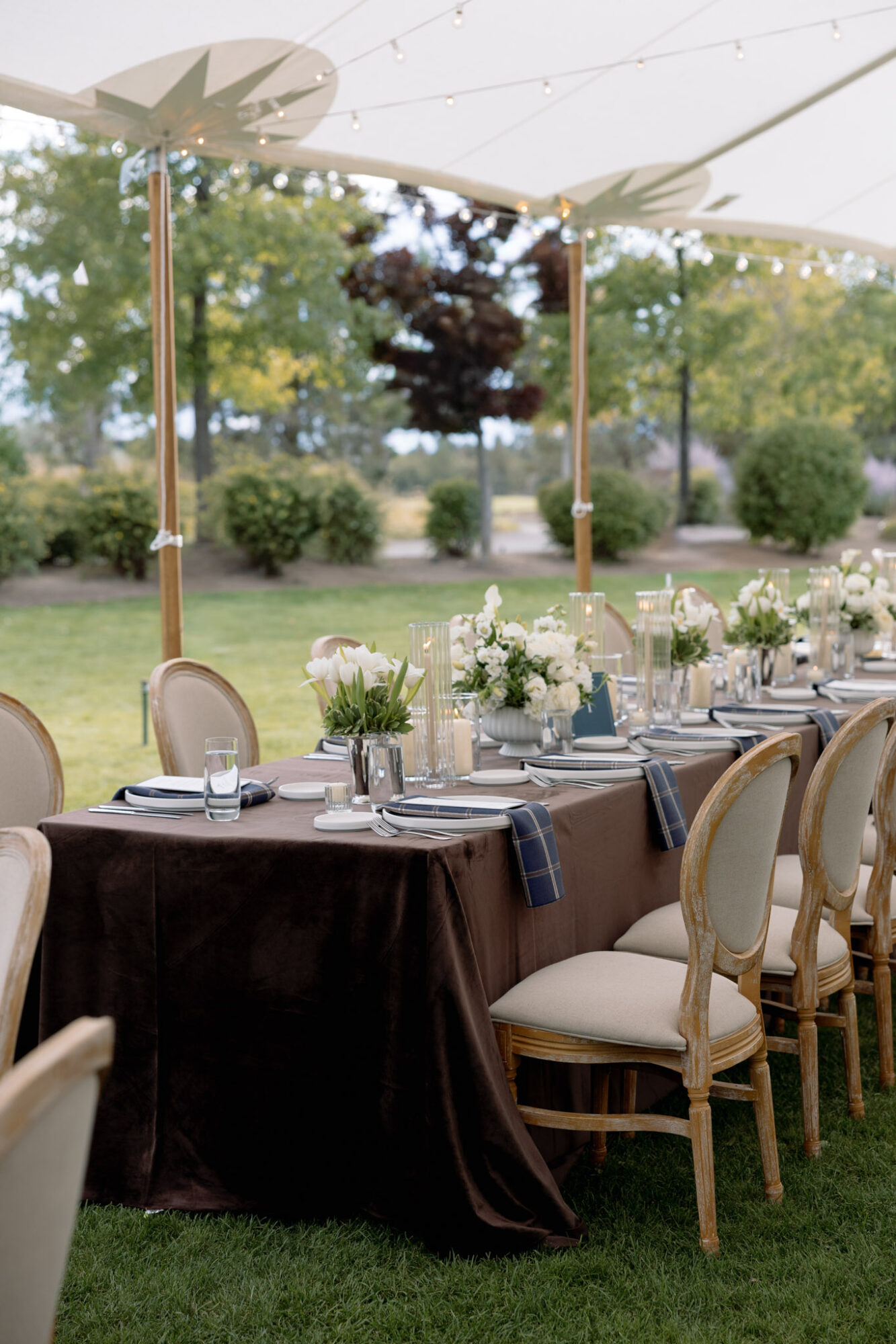 Outdoor dining table with white flowers, set under a large umbrella, surrounded by chairs, in a garden setting.