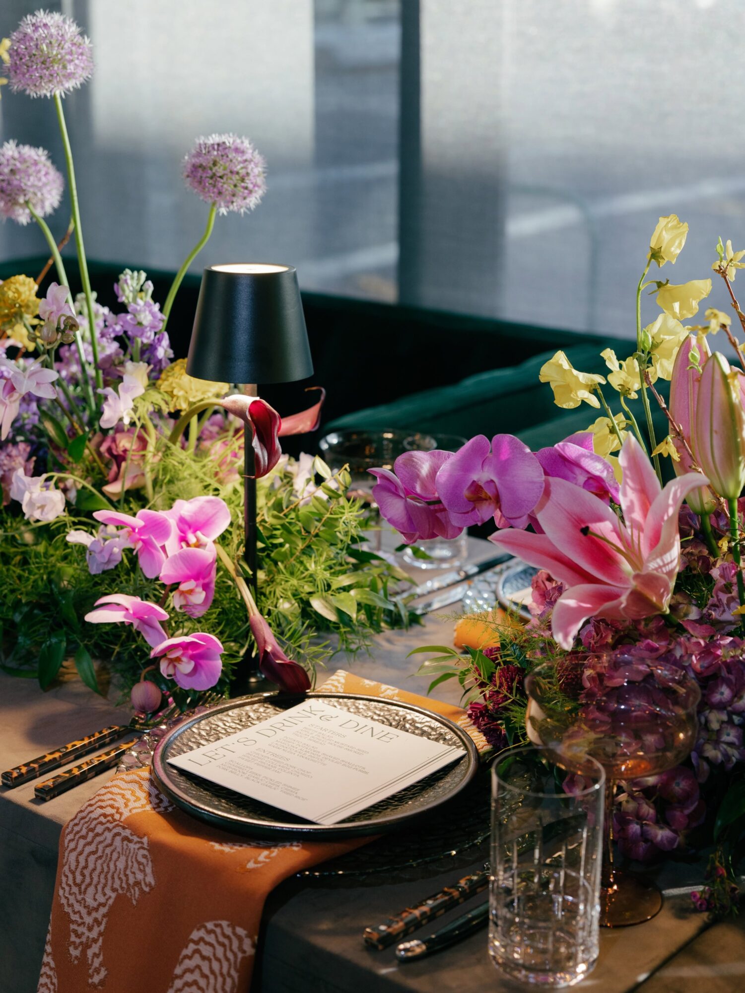 Table with floral arrangements, a black lamp, a glass, and a notebook, near a window with blinds.
