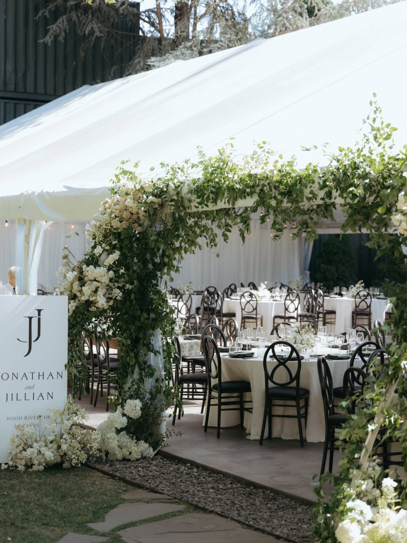 Outdoor event setup with tables, chairs, and floral decorations under a white canopy, greenery, and sunlight.