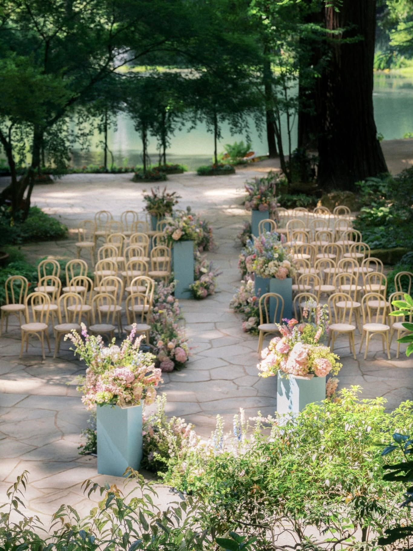 Outdoor wedding setup with rows of chairs, floral arrangements, and trees in a park-like setting.