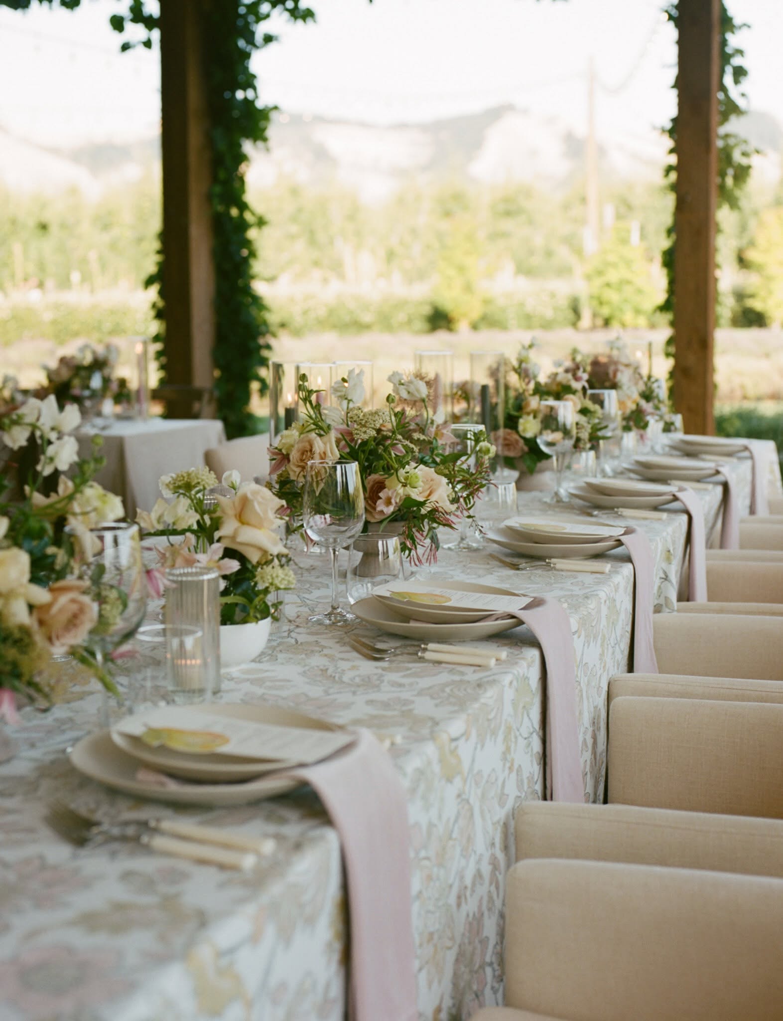 Long dining table with floral arrangements, plates, glasses, and chairs in a bright outdoor setting.