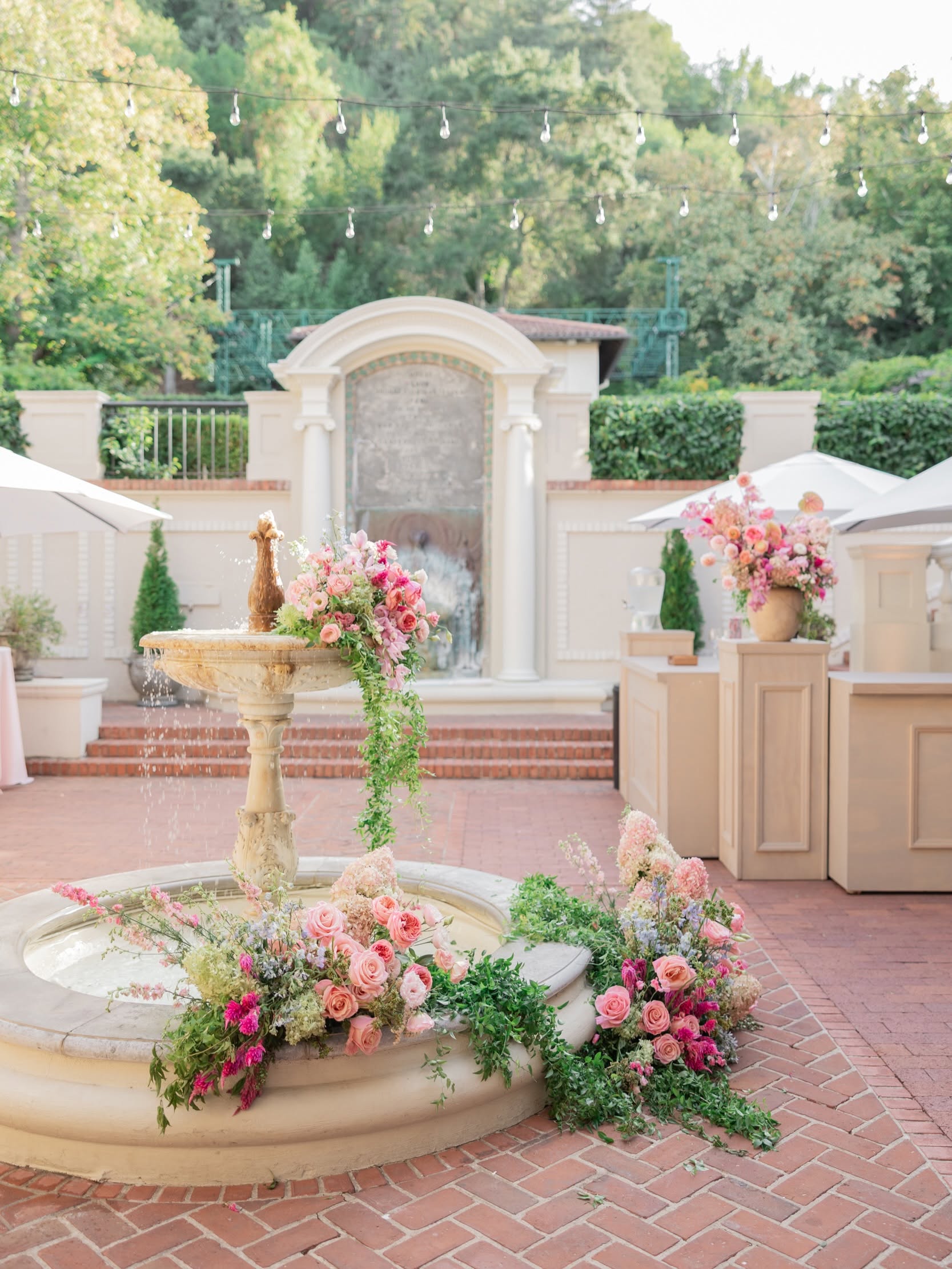 Decorative fountain surrounded by pink and white flowers on a brick pathway, with a stone monument and greenery in background.