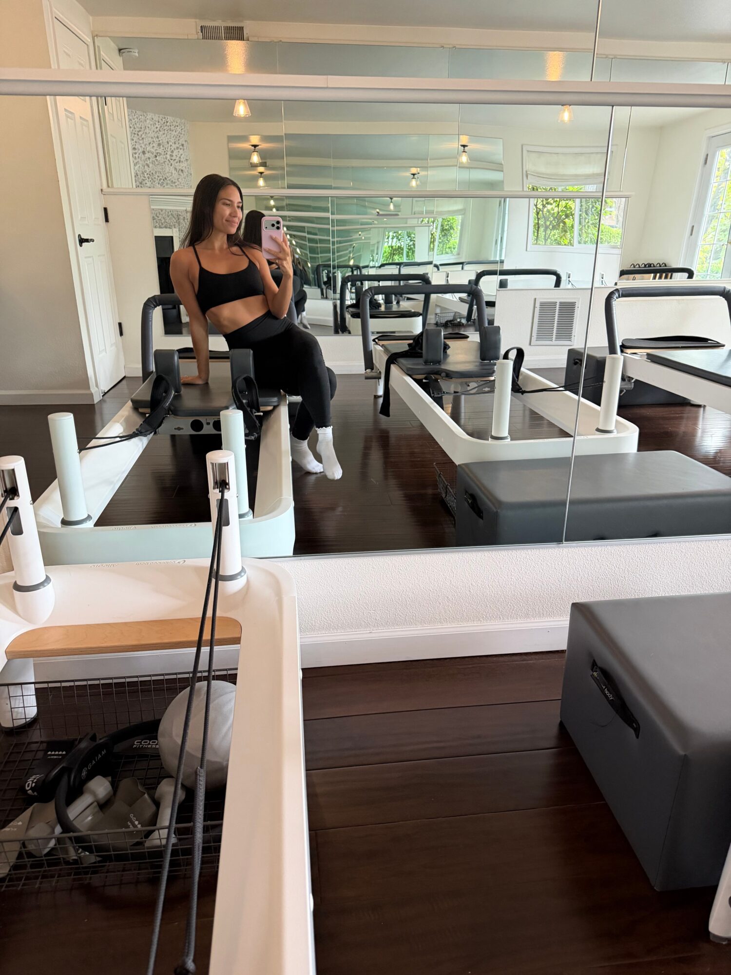 A woman in a gym taking a mirror selfie, with exercise equipment and large windows in the background.