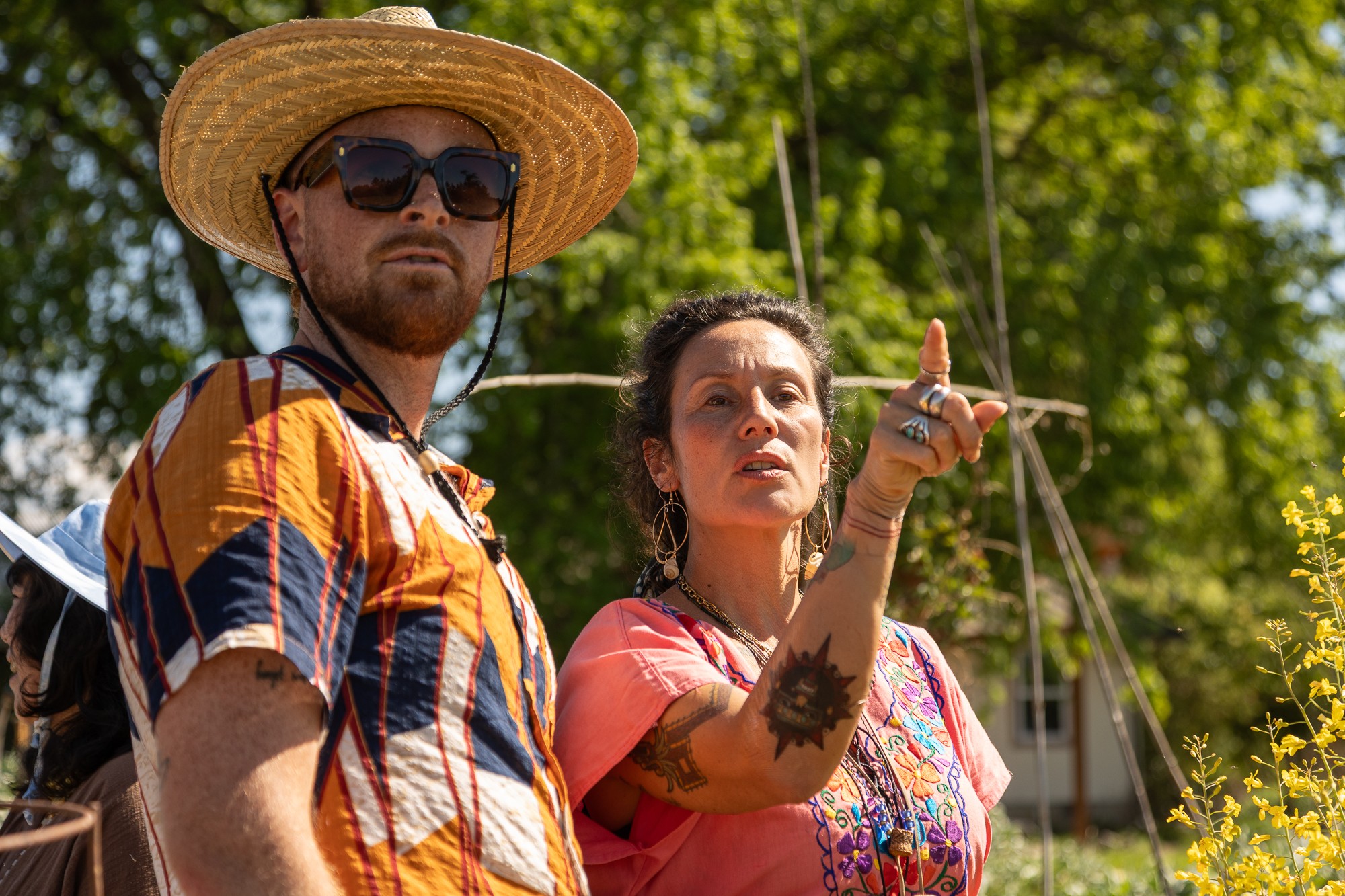 Two people outdoors, one wearing a large straw hat and sunglasses, the other pointing with a serious expression, trees in background.