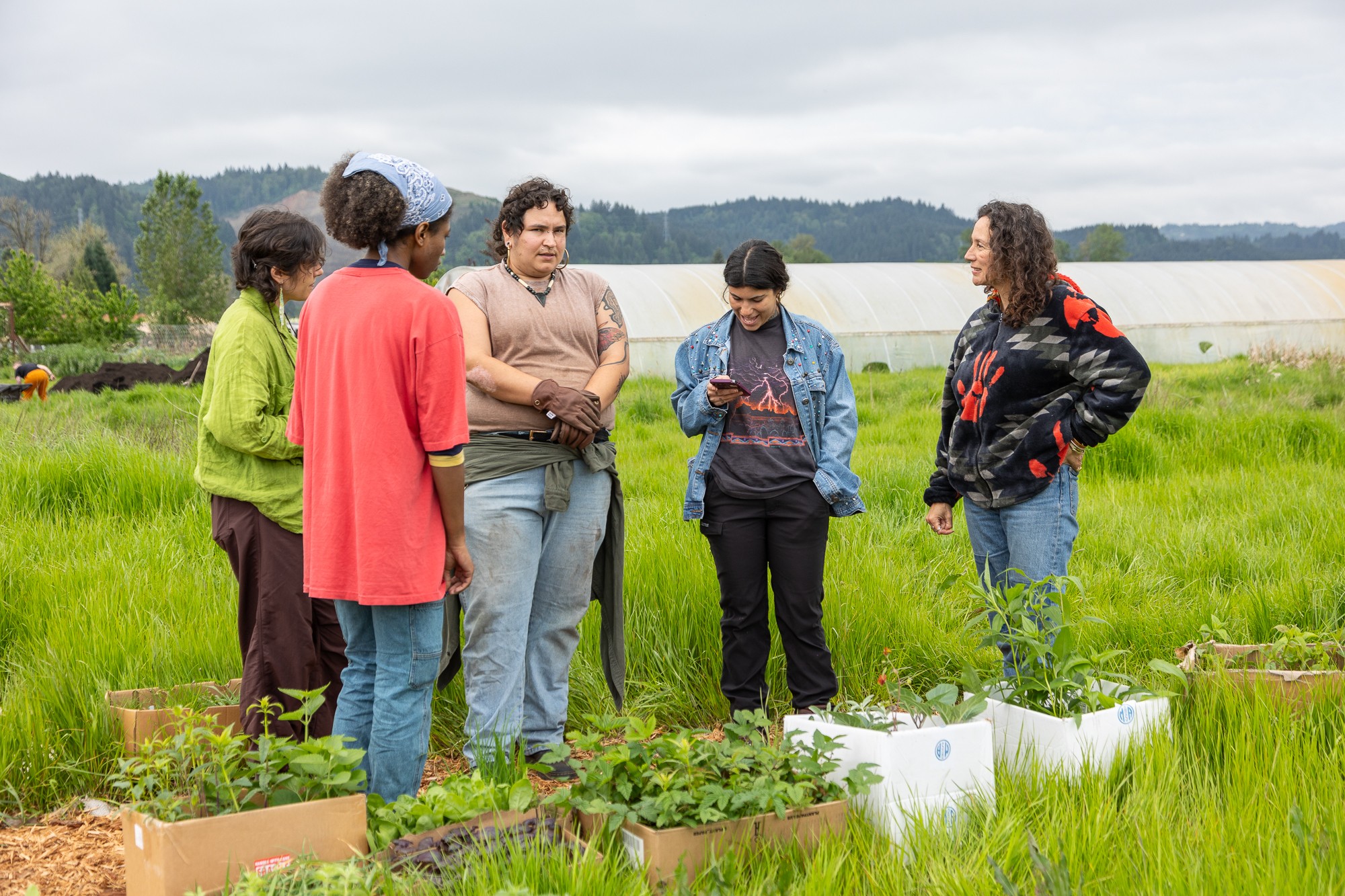Group of six people standing outdoors in a green field, engaged in conversation, with plants and a greenhouse in the background.