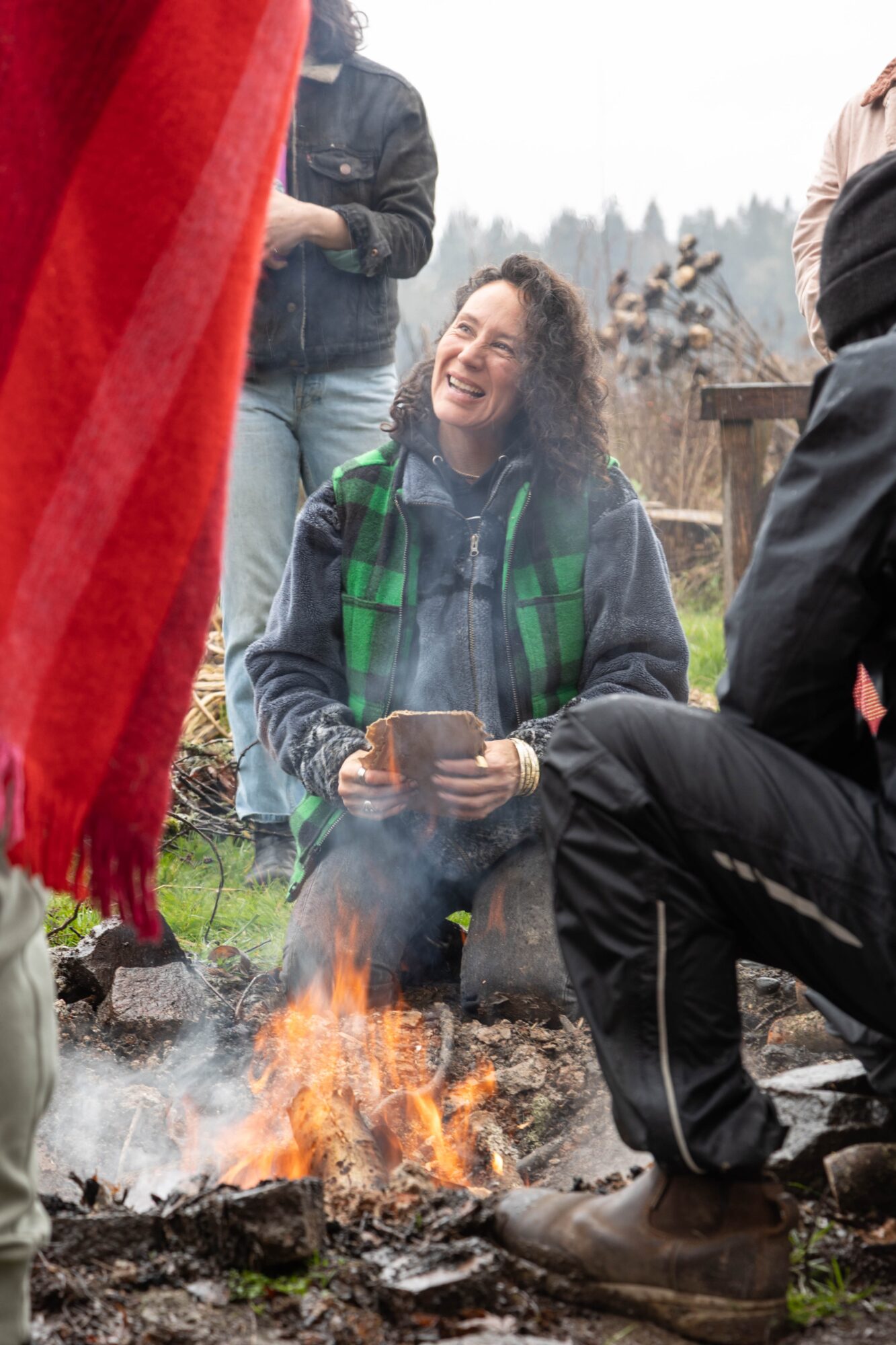 Woman sitting outdoors near a campfire, smiling, holding a cup, surrounded by people, with trees and sky in background.