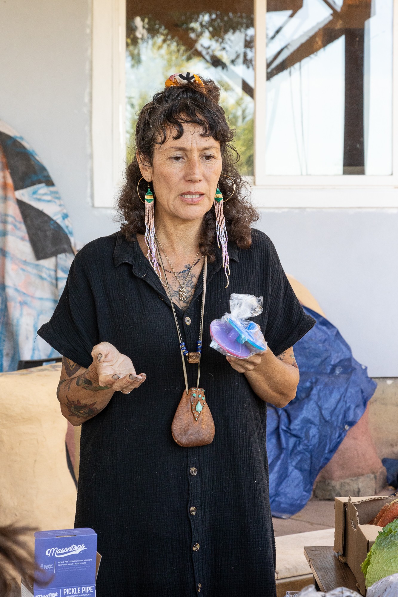 Woman with curly hair and earrings holding a phone and a small object, standing outdoors near a window and a blue bag.