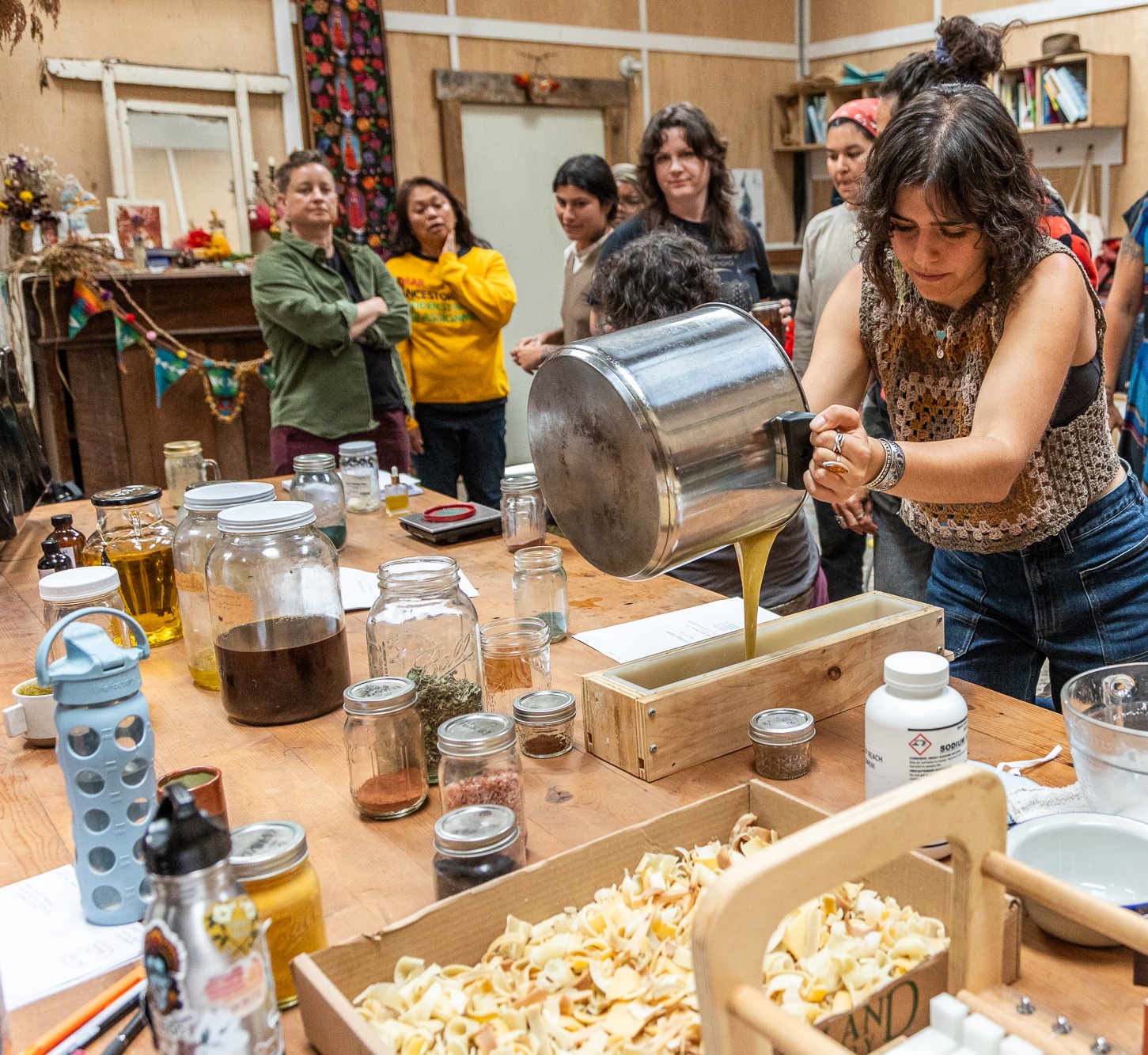 A woman pours liquid into a box while others watch in a room with shelves and decorations, cluttered table with jars and snacks.