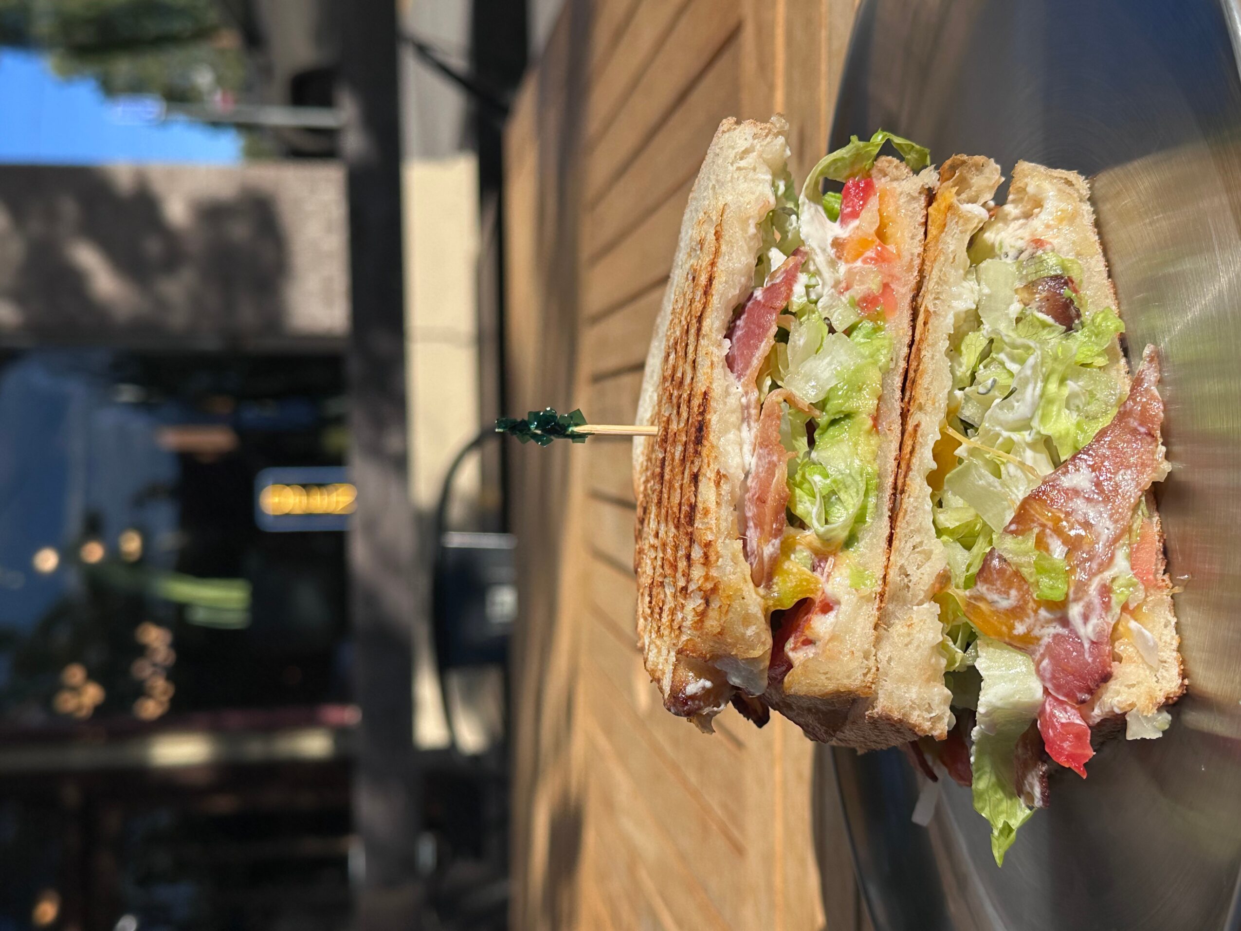 Half-eaten sandwich with lettuce, tomato, and meat on toasted bread, on a metal tray outdoors.