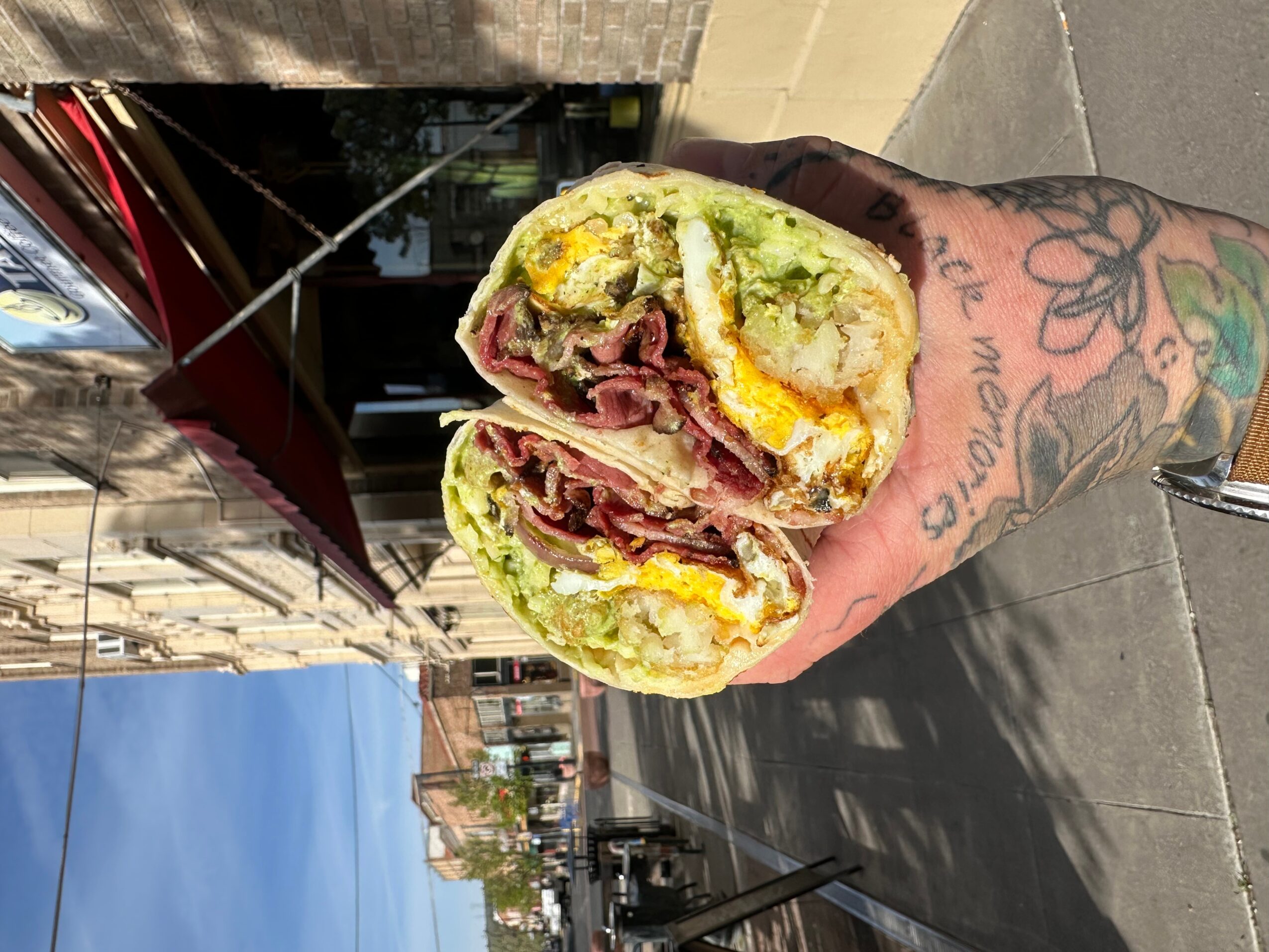 Person holding a sandwich with lettuce, meat, cheese, and other ingredients, outdoors on a sidewalk.