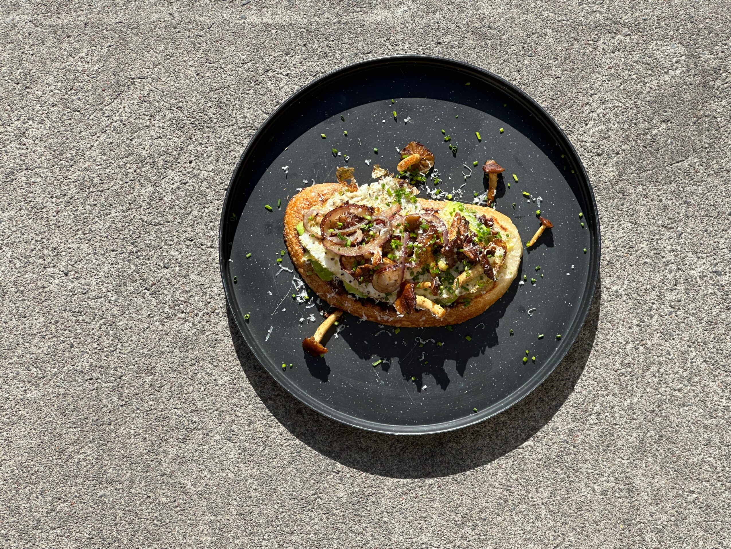 Half avocado with toppings on a black plate, on a gray surface, with scattered crumbs and garnishes.