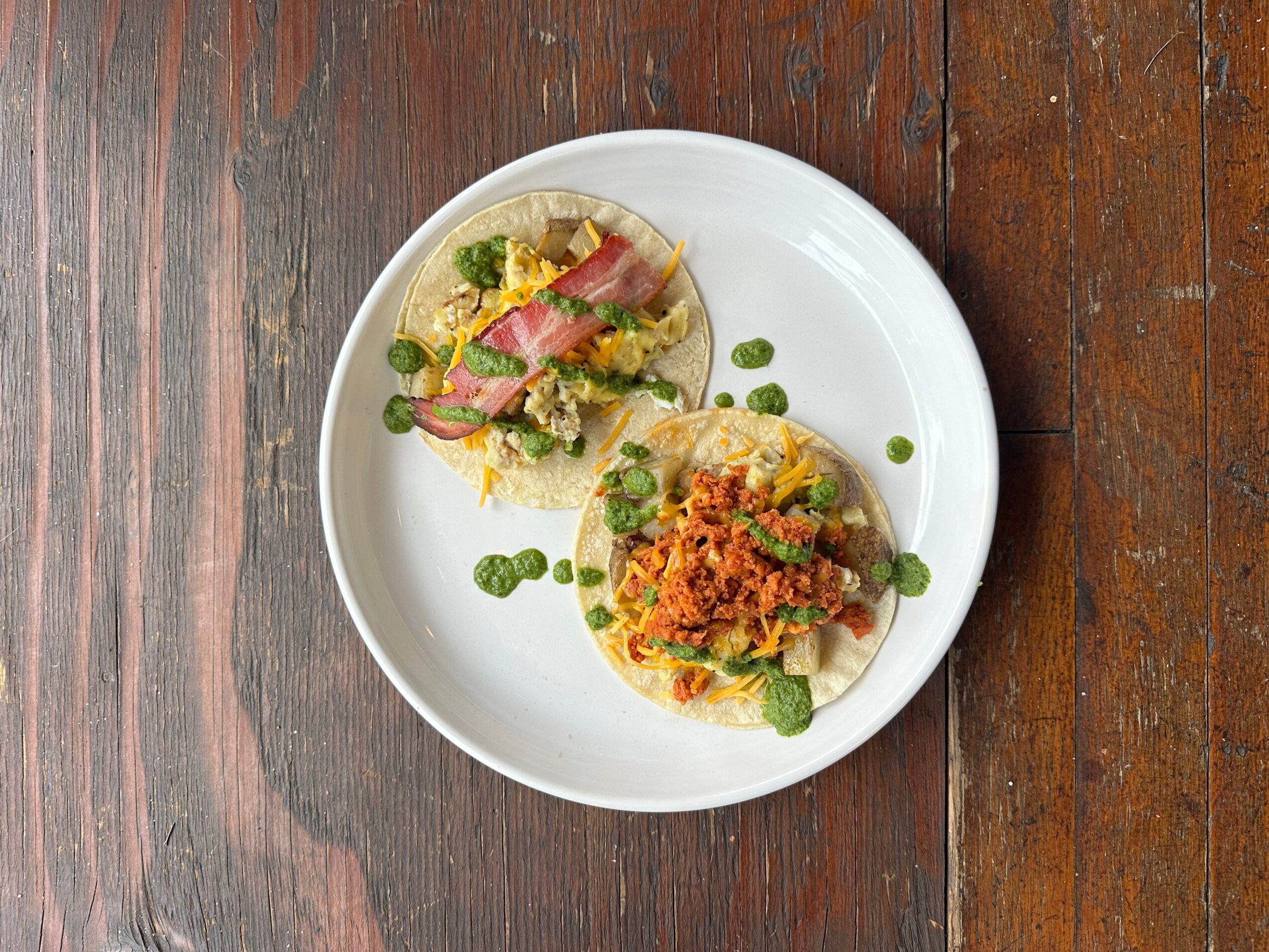 Two tacos on a white plate with green sauce drops, on a wooden table.