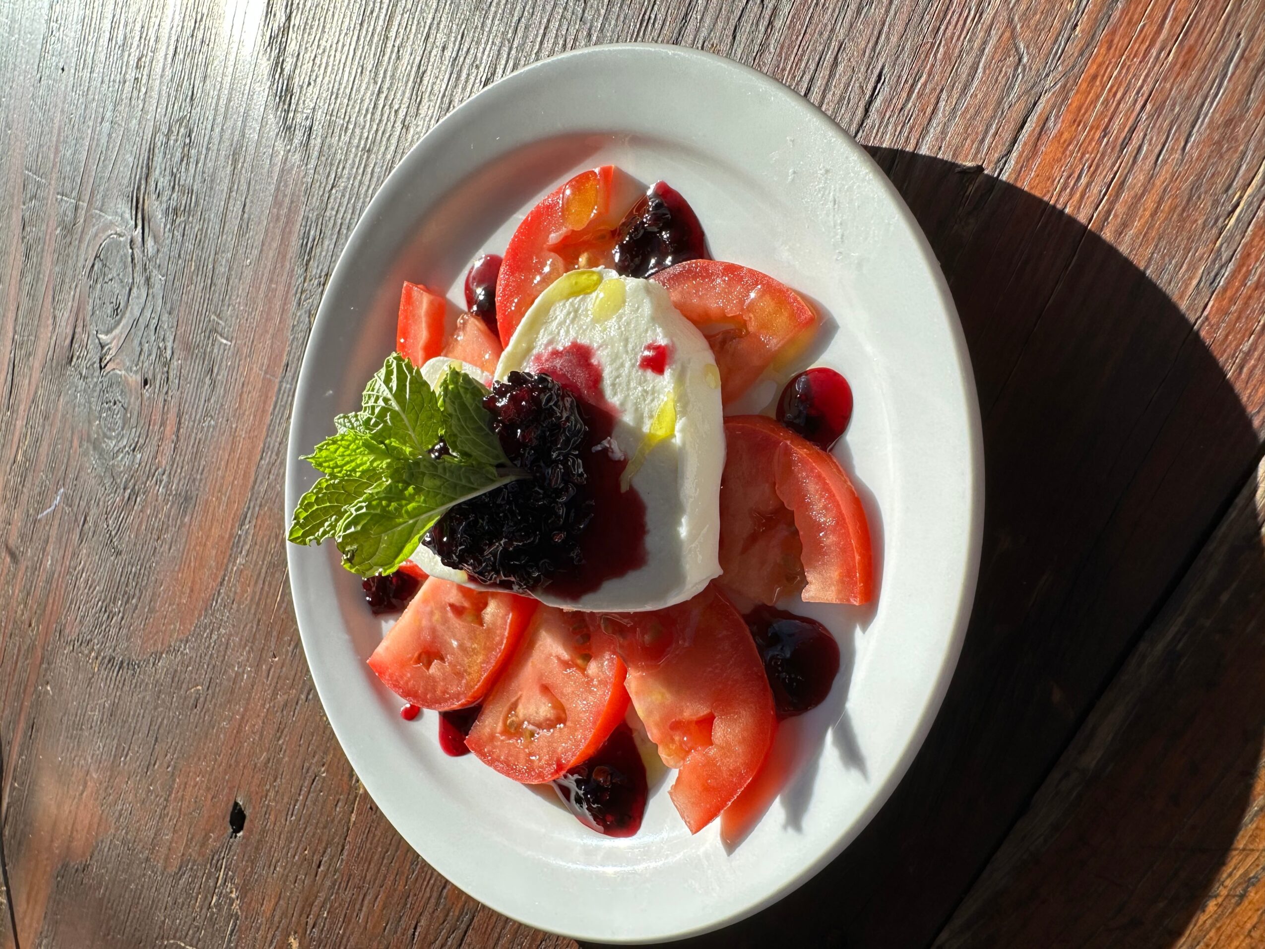 Bowl of mixed fresh fruit with a dollop of cream and mint leaf, on a wooden surface, sunlight shining.