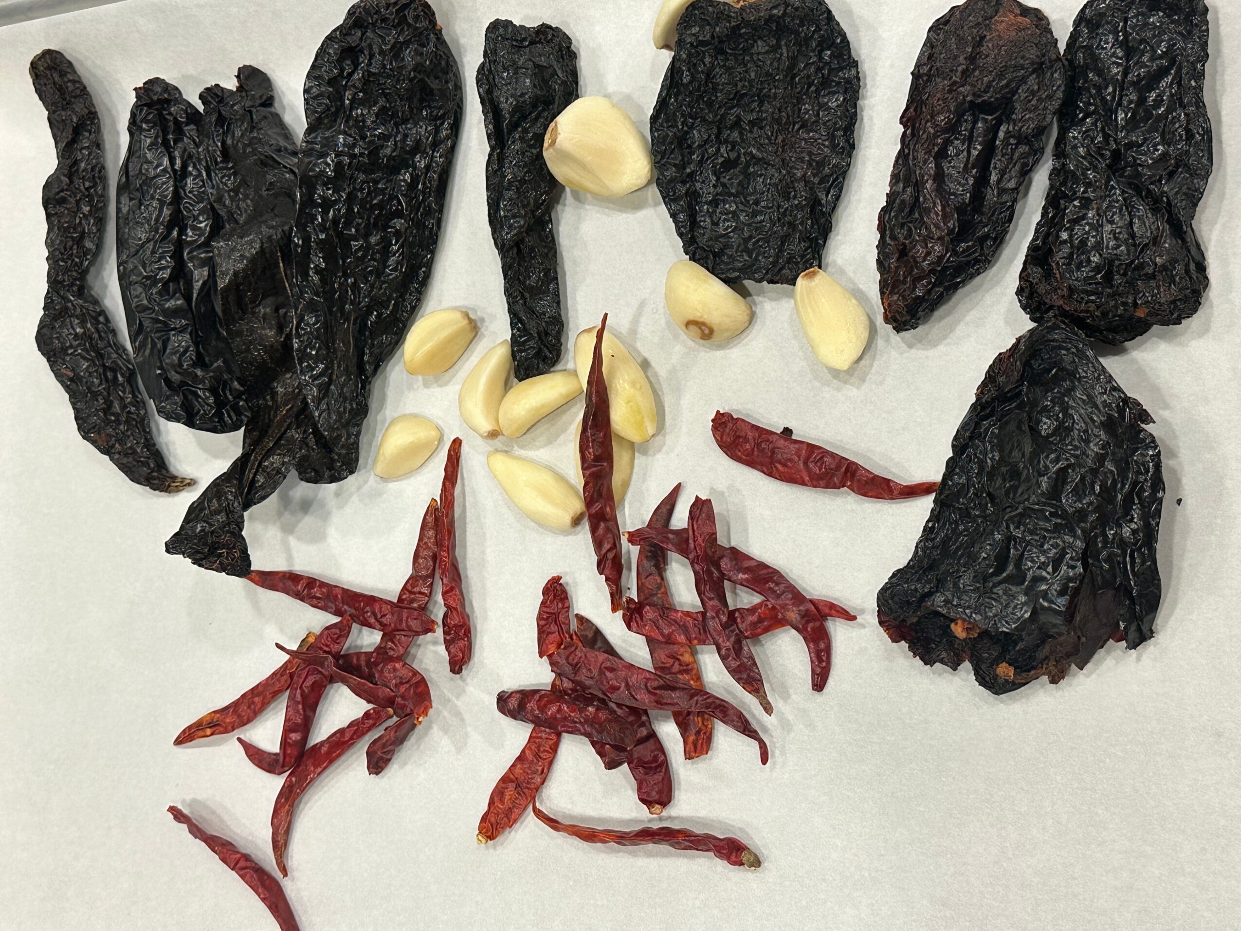 Dried black chili peppers, garlic cloves, and dried red chili peppers on a white surface.