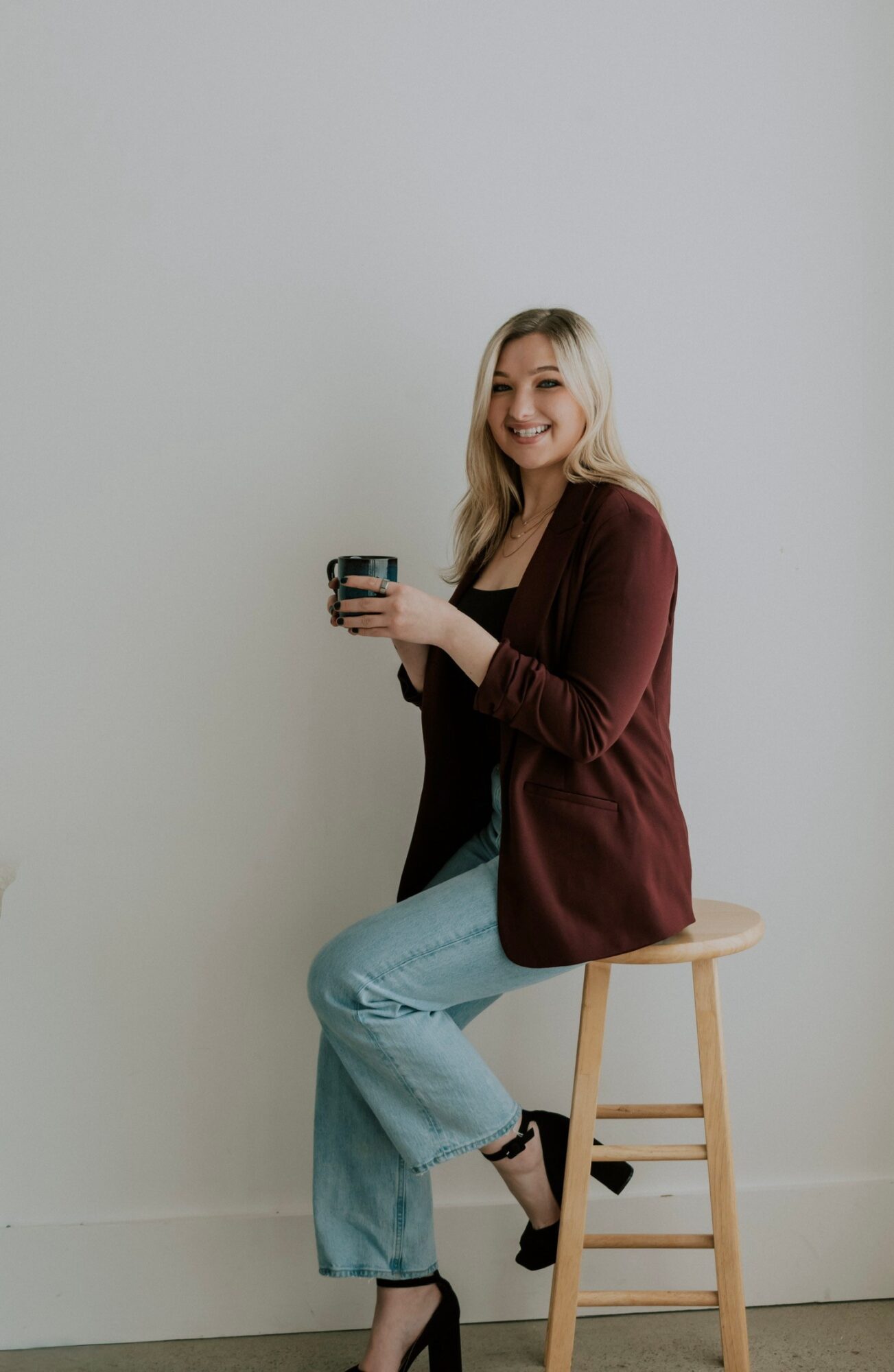 Woman sitting on a stool, smiling, holding a cup, wearing a maroon cardigan and light blue jeans, against a plain wall.