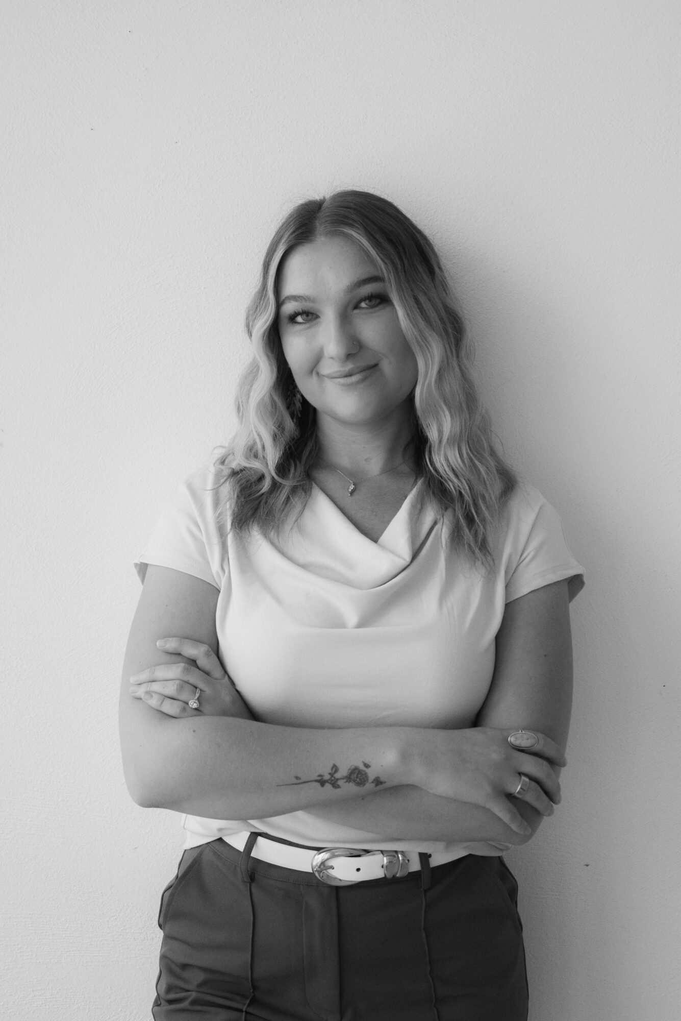 Young woman with wavy hair smiling, arms crossed, wearing a short-sleeved top and dark pants, standing against a plain wall.
